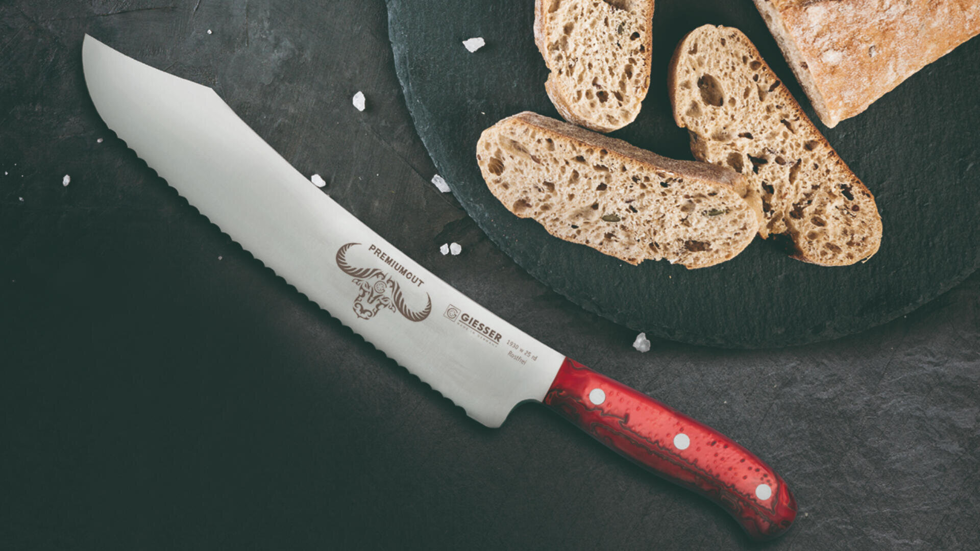 Ein gezacktes Brotmesser mit rotem Griff liegt auf einer dunklen Fläche neben rustikalen Brotscheiben auf einer runden Schieferplatte, daneben ist grobes Salz gestreut.