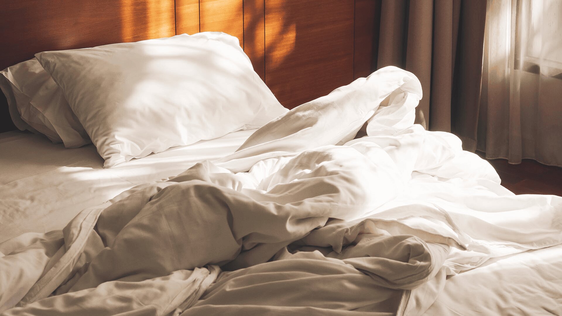 Ein Bett mit weißen Kissen und zerknitterten weißen Laken ist in warmes Sonnenlicht getaucht, das durch ein nahe gelegenes Fenster fällt und weiche Schatten auf die zerknitterte Bettwäsche wirft.