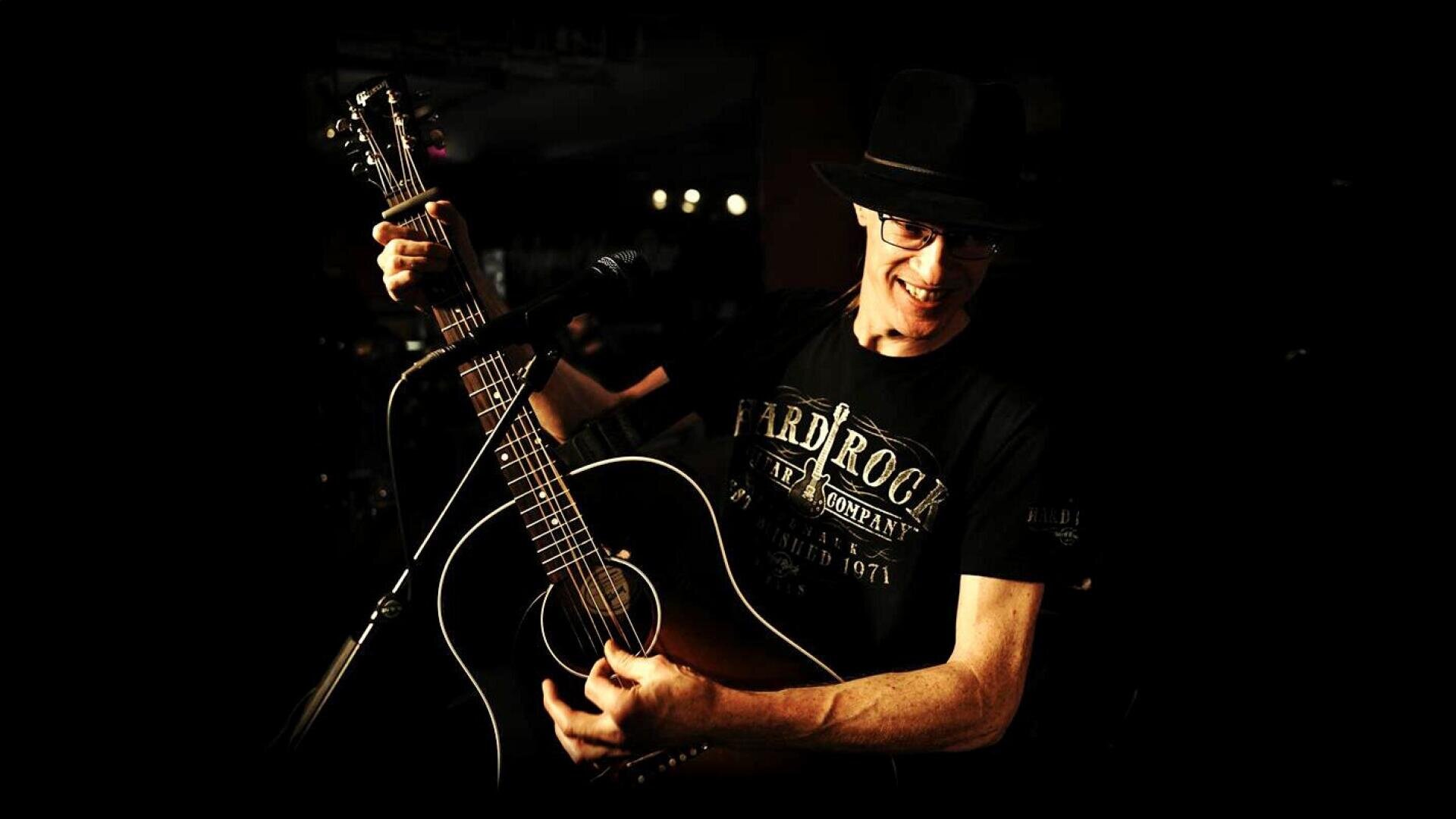 Ein lächelnder Musiker mit Brille und schwarzem Hut spielt bei dramatischer Beleuchtung energisch auf einer akustischen Gitarre. Er trägt ein schwarzes Hard Rock Cafe-T-Shirt, und der Hintergrund ist dunkel und unscharf.