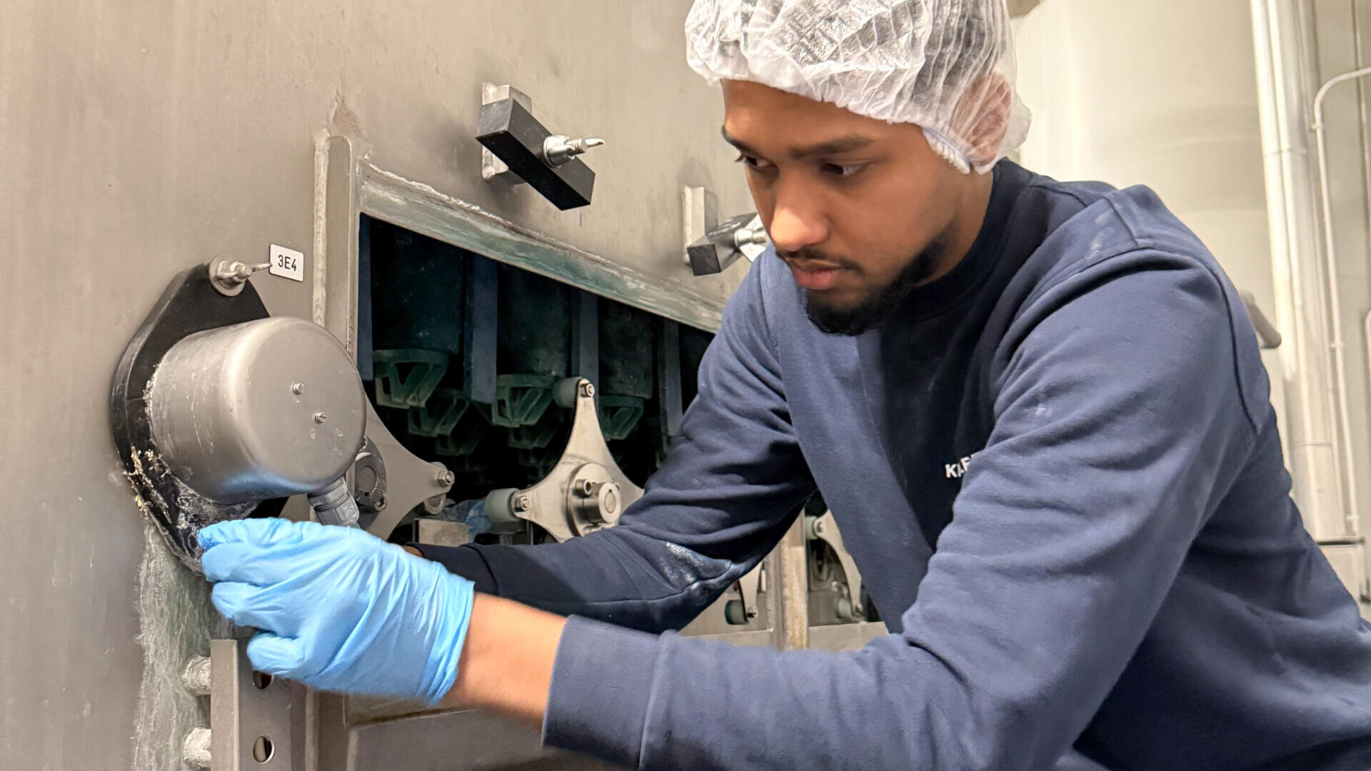 Eine Person, die ein Haarnetz, blaue Handschuhe und ein marineblaues Sweatshirt trägt, arbeitet an einer großen Industriemaschine und justiert oder prüft ein Metallteil in einer Fabrik oder einem Verarbeitungsbetrieb.