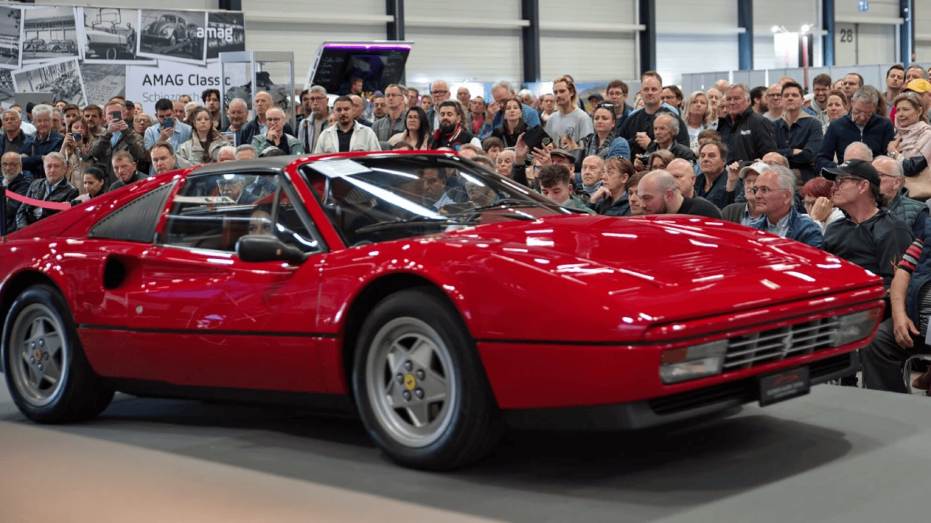 Ein klassischer roter Ferrari-Sportwagen wird in einem Innenbereich ausgestellt, umgeben von einer großen Menschenmenge, die das Auto bei einer Automobilveranstaltung oder -ausstellung aufmerksam beobachtet und fotografiert.