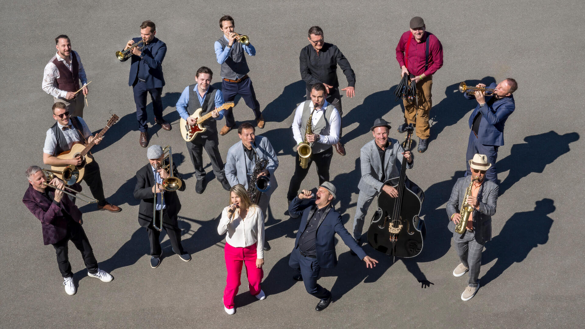 Eine Gruppe von vierzehn Musikern, meist Männer und eine Frau, steht in einem losen Kreis auf einer gepflasterten Fläche und spielt verschiedene Instrumente, darunter Blechblasinstrumente, Gitarren und Saxophone. Sie scheinen enthusiastisch zu spielen.
