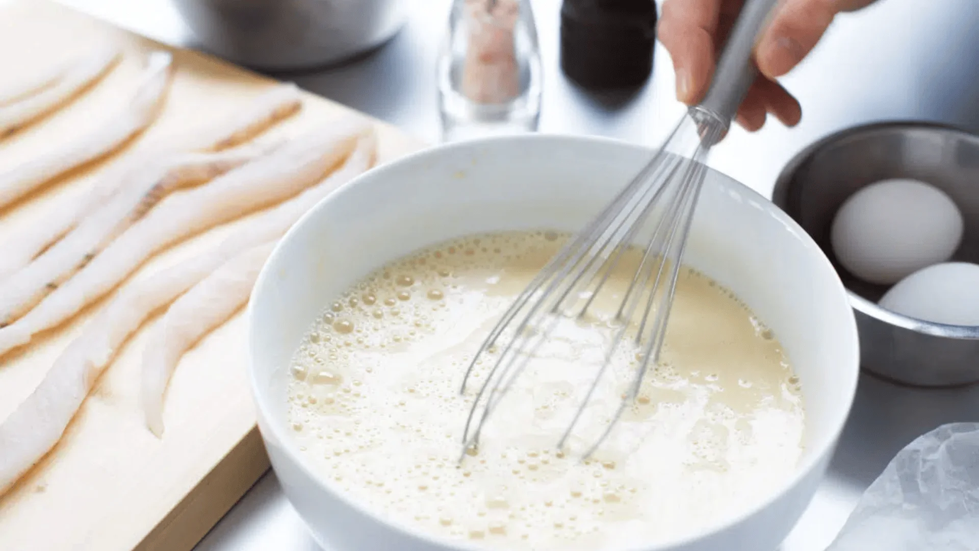 Eine Hand rührt mit einem Schneebesen einen cremigen Teig in einer weißen Schüssel, während rohe Fischfilets auf einem Holzbrett daneben liegen und Eier und Küchenzutaten im Hintergrund zu sehen sind.