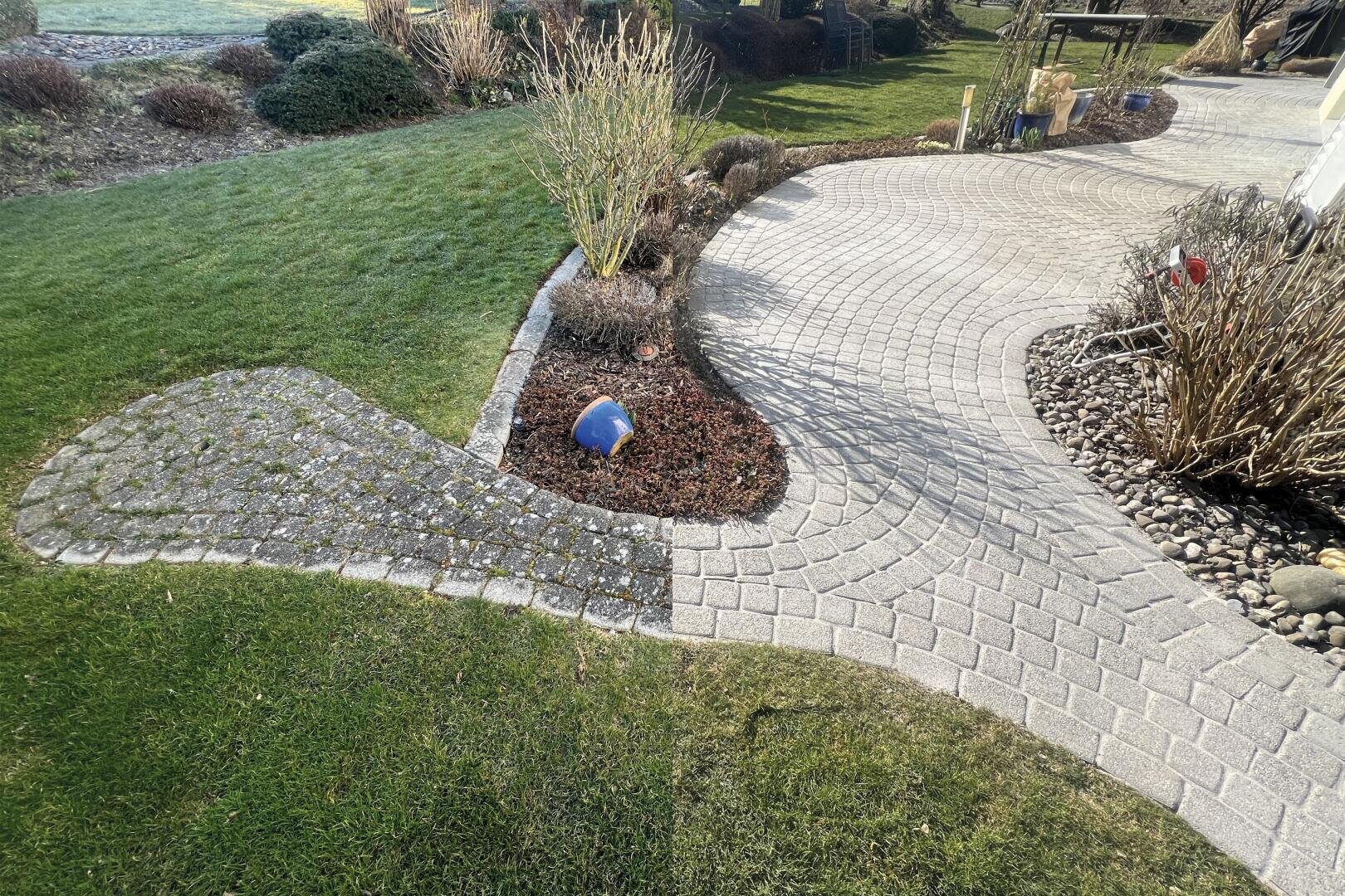 Ein geschwungener Steinweg, der von Gras und Gartenbeeten mit kahlen Sträuchern gesäumt wird. Ein kleines blaues und rotes Spielzeug ist teilweise im Mulch am Wegrand vergraben. Die Szene ist gut beleuchtet, was auf einen sonnigen Tag in einem Landschaftsgarten hindeutet.