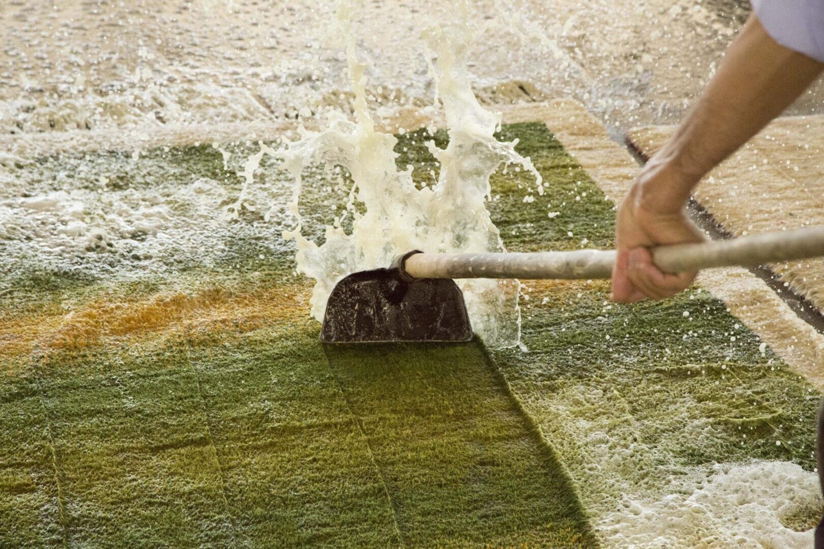 Eine Person schrubbt mit einem Werkzeug mit langem Stiel einen seifigen Teppich, so dass während des Reinigungsvorgangs Wasser und Schaum hochspritzen.