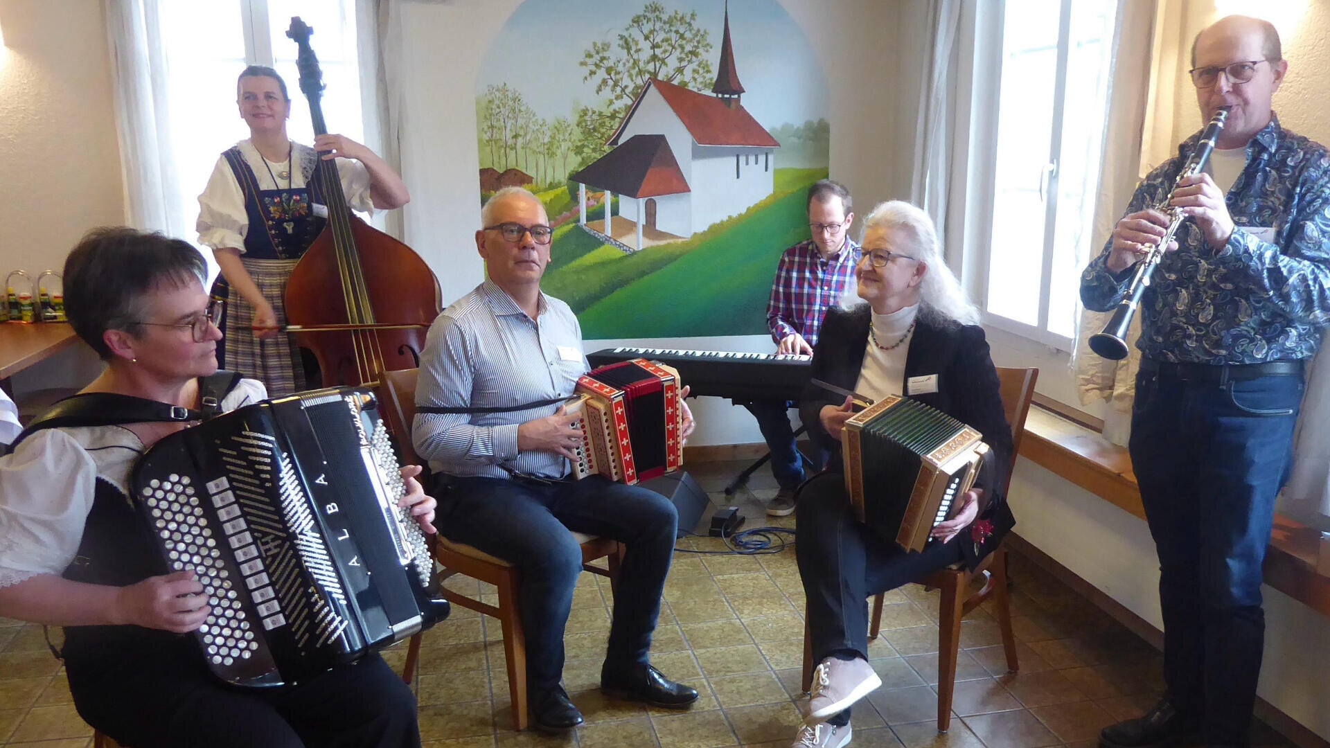Sechs Musiker spielen drinnen gemeinsam auf traditionellen Instrumenten; zwei spielen Akkordeon, einer auf einem kleinen Akkordeon, einer auf dem Kontrabass, einer auf der Klarinette und einer auf dem Keyboard. An der Wand hinter ihnen ist eine Wandmalerei einer Kirche zu sehen.