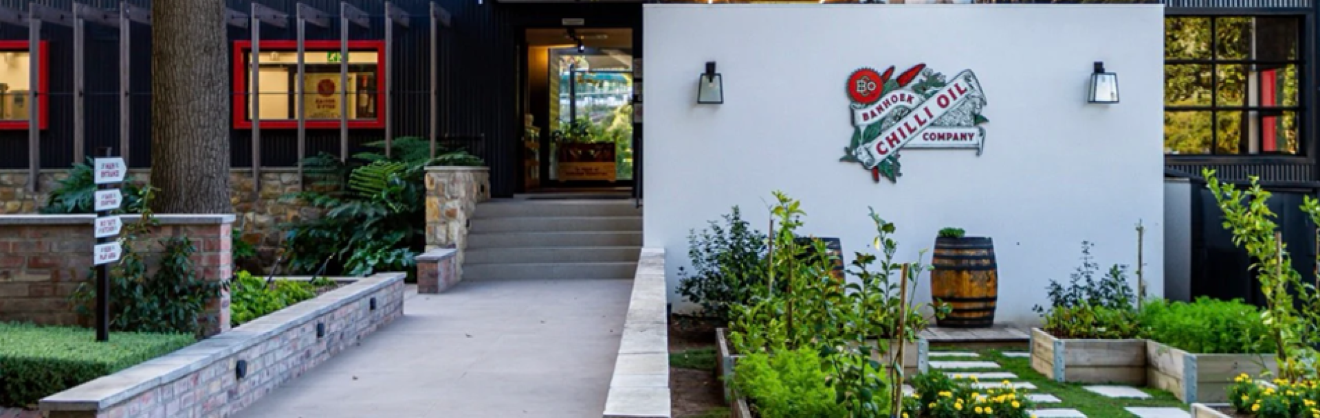 Eingang zu einem Gebäude mit einer weißen Wand, auf der das Logo der "Brooklyn Chilli Oil Company" prangt, umgeben von Grünpflanzen, einem kleinen Garten, Fässern und großen Fenstern mit roten Rahmen.