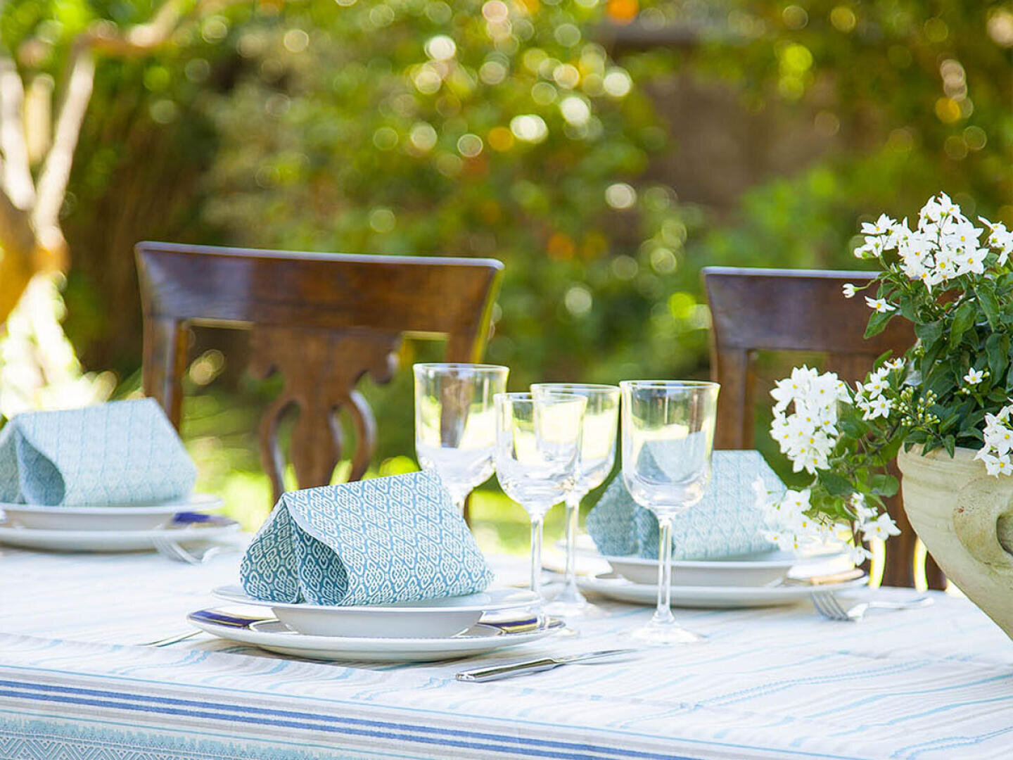 Ein sonnenbeschienener Tisch im Freien ist mit gemusterten blauen Servietten, weißen Tellern, Gläsern und einer Vase mit weißen Blumen gedeckt, während im Hintergrund Holzstühle und Grünpflanzen zu sehen sind.