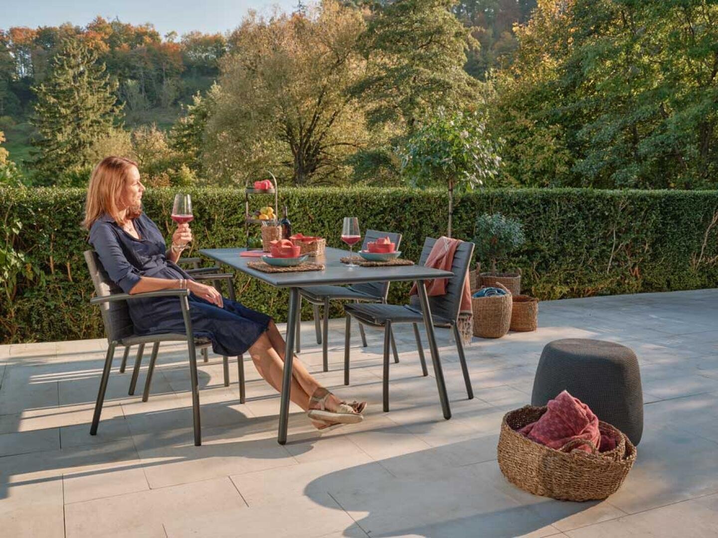Eine Frau in einem marineblauen Kleid sitzt mit einem Glas Wein in der Hand an einem Esstisch im Freien auf einer Veranda. Der Tisch ist für zwei Personen gedeckt, umgeben von Grünpflanzen und Bäumen, mit Körben und Kissen in der Nähe.