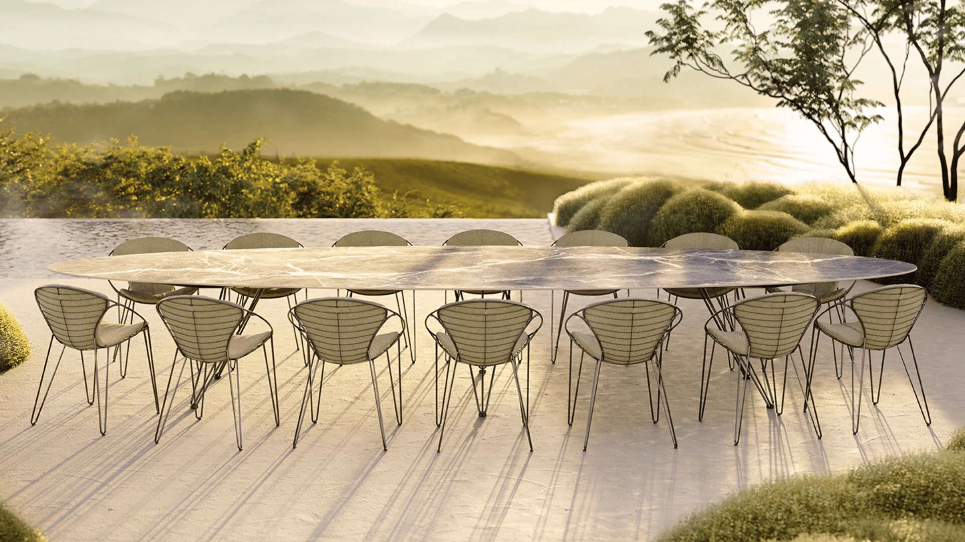 Ein langer, eleganter Esstisch im Freien mit fünfzehn modernen Drahtstühlen steht auf einer Terrasse mit Blick auf sanfte grüne Hügel und eine neblige Landschaft im weichen Sonnenlicht.