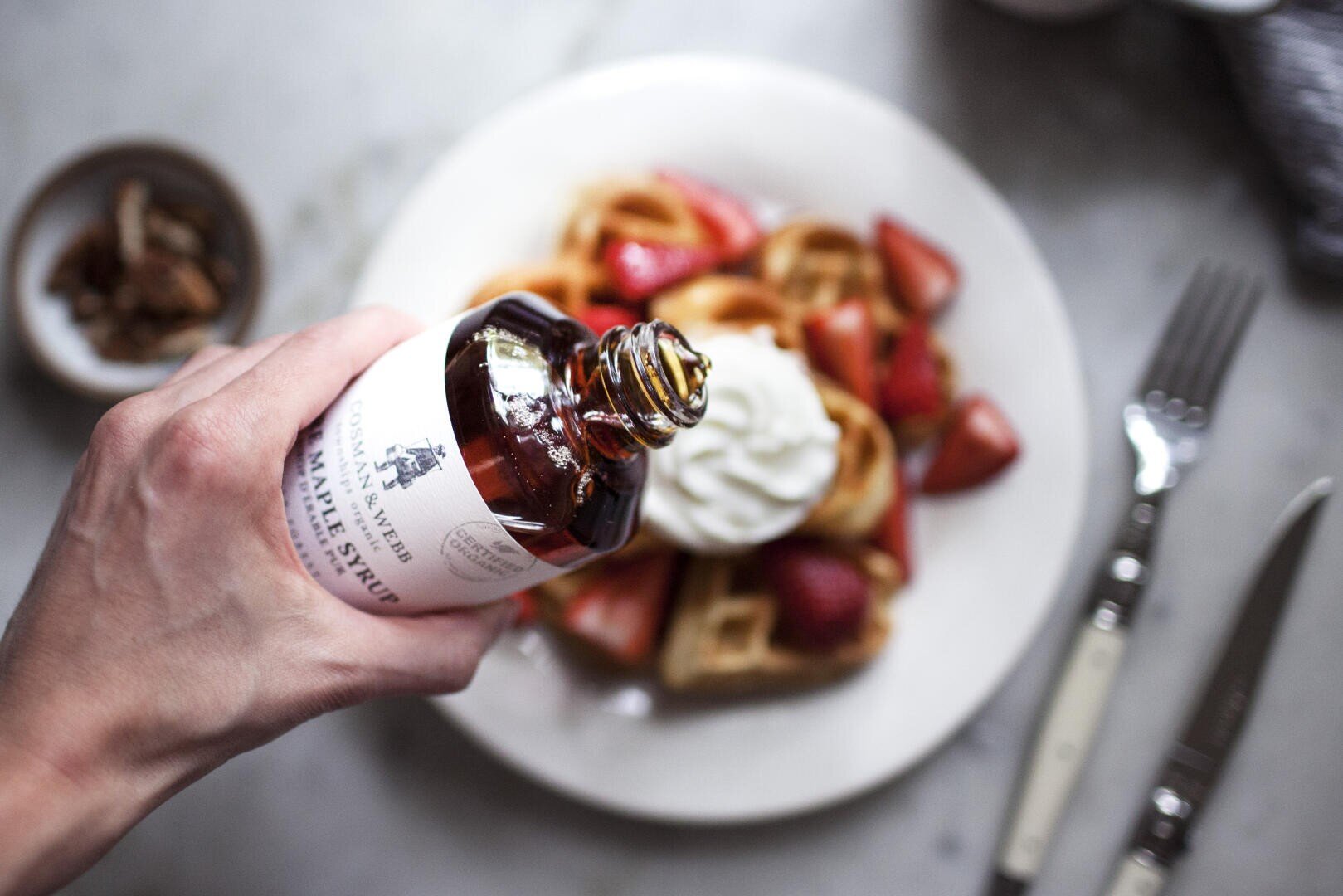 Eine Hand gießt Ahornsirup aus einer Flasche auf die Waffeln, die mit Schlagsahne und frischen Erdbeeren auf einem weißen Teller liegen, Gabel und Messer liegen daneben.