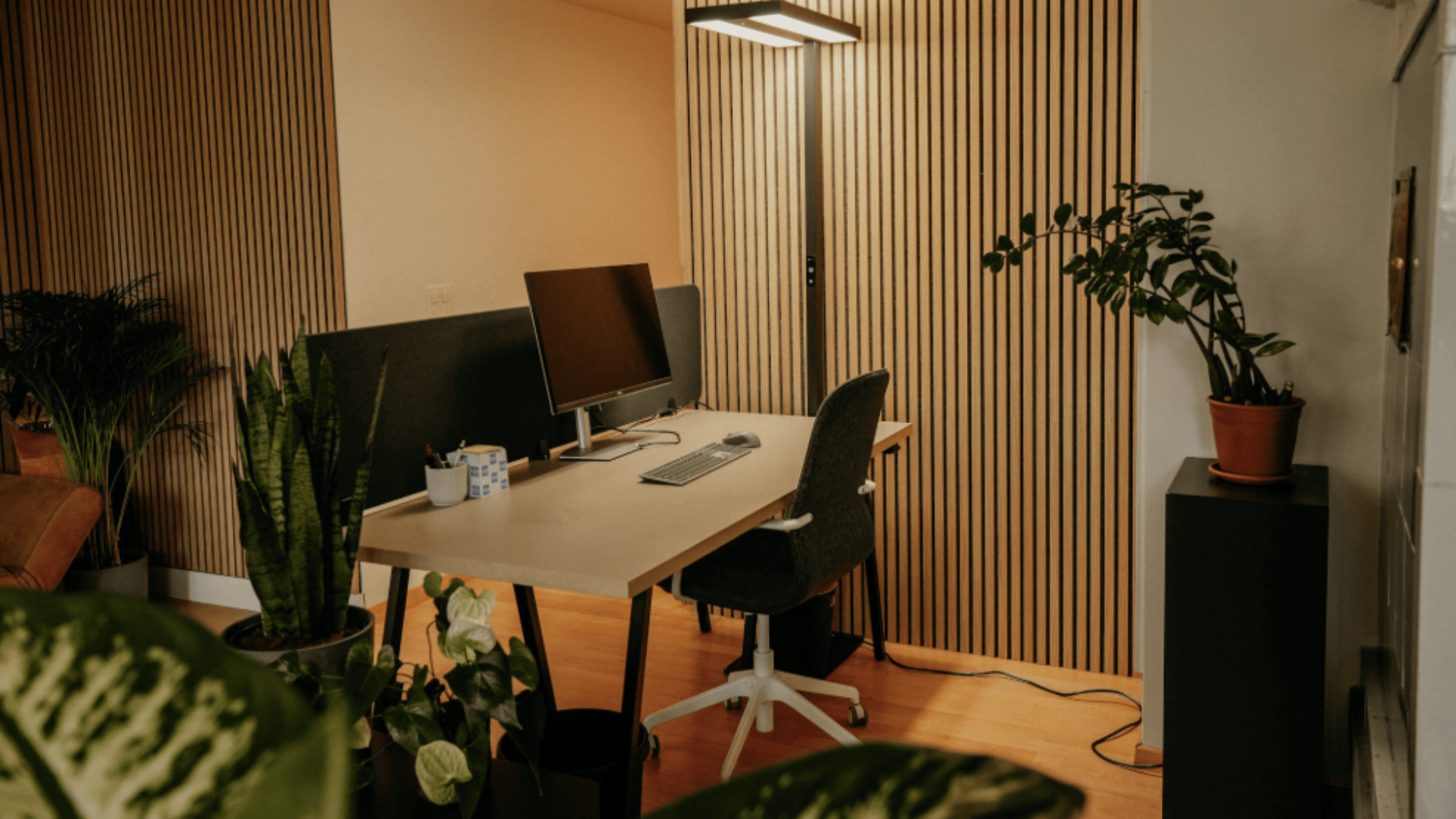 Ein modernes Heimbüro mit einem Holzschreibtisch, Computermonitor, Tastatur und Bürostuhl. Der Raum ist mit Holzlattenwänden, Topfpflanzen und warmer Beleuchtung ausgestattet, die eine gemütliche und einladende Atmosphäre schaffen.