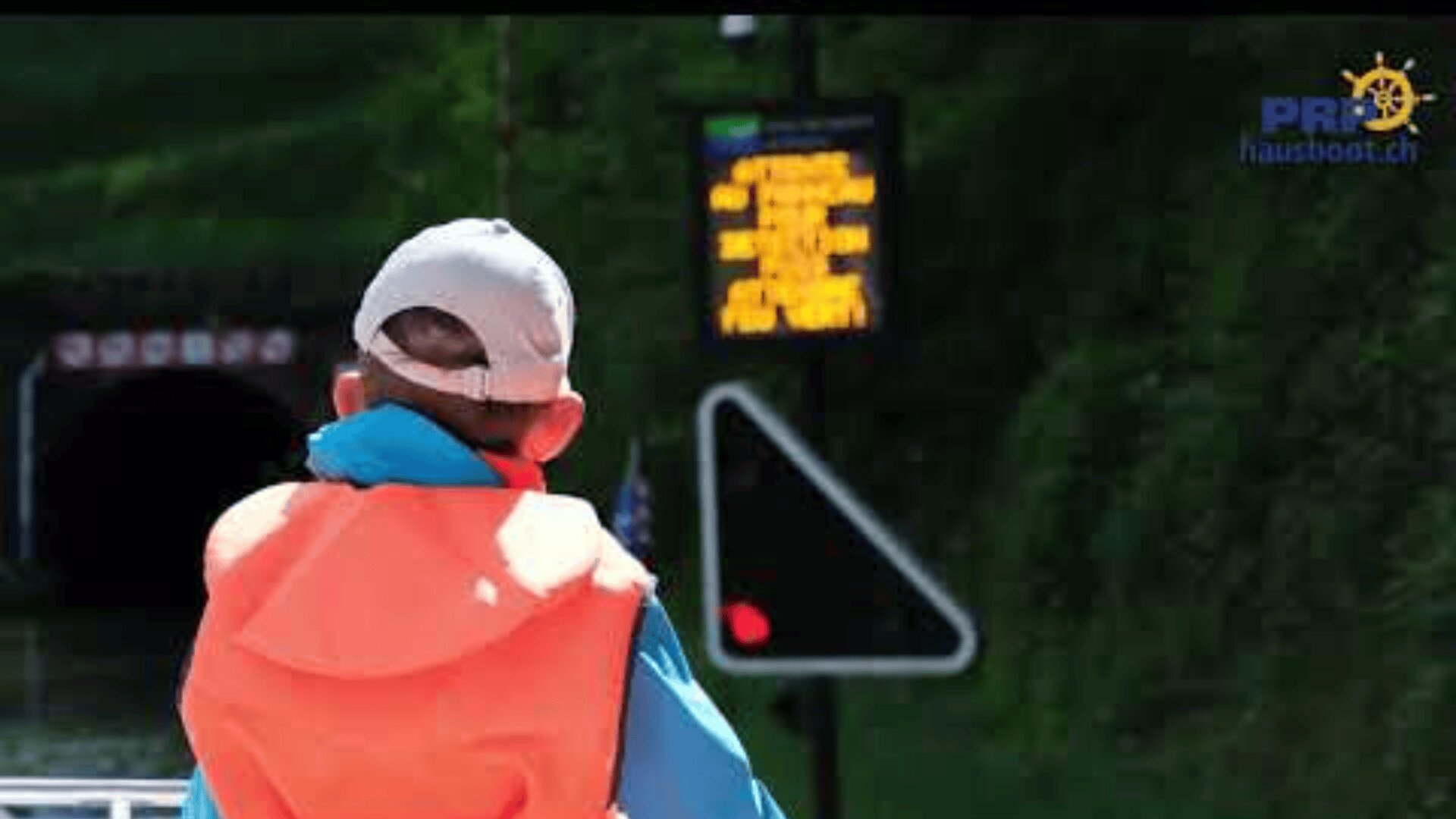 Eine Person, die eine helle Mütze, eine blaue Jacke und eine orangefarbene Schwimmweste trägt, steht vor einem elektronischen Schild in der Nähe eines Tunneleingangs, mit Grünzeug im Hintergrund und einem Logo in der oberen rechten Ecke.