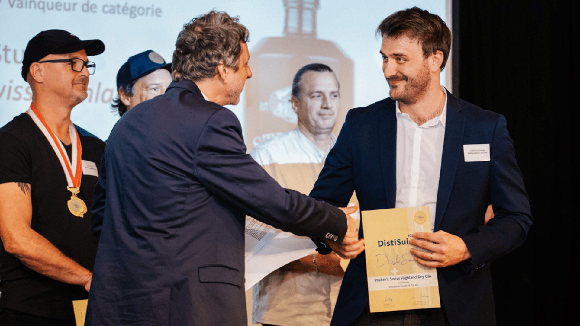 Ein Mann im Anzug schüttelt einem anderen Mann, der eine Urkunde hält, bei einer Preisverleihung die Hand. Zwei weitere Männer, von denen einer eine Medaille trägt, stehen im Hintergrund, während hinter ihnen ein Bildschirm mit Text angezeigt wird.