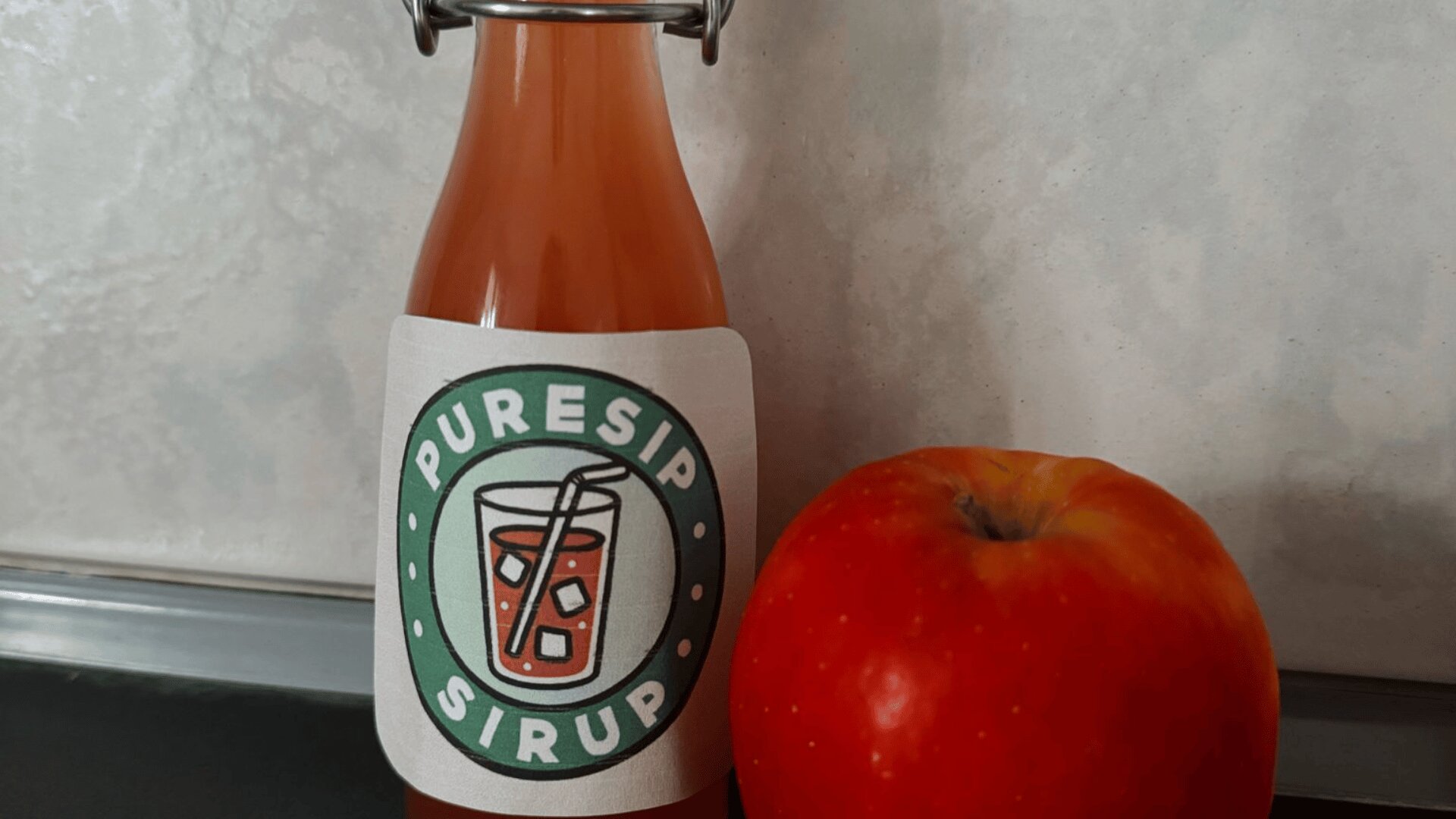 Eine Flasche mit der Aufschrift PureSip Sirup mit einer Getränkegrafik, die neben einem roten Apfel auf einer dunklen Fläche vor einem hellen, strukturierten Hintergrund steht.