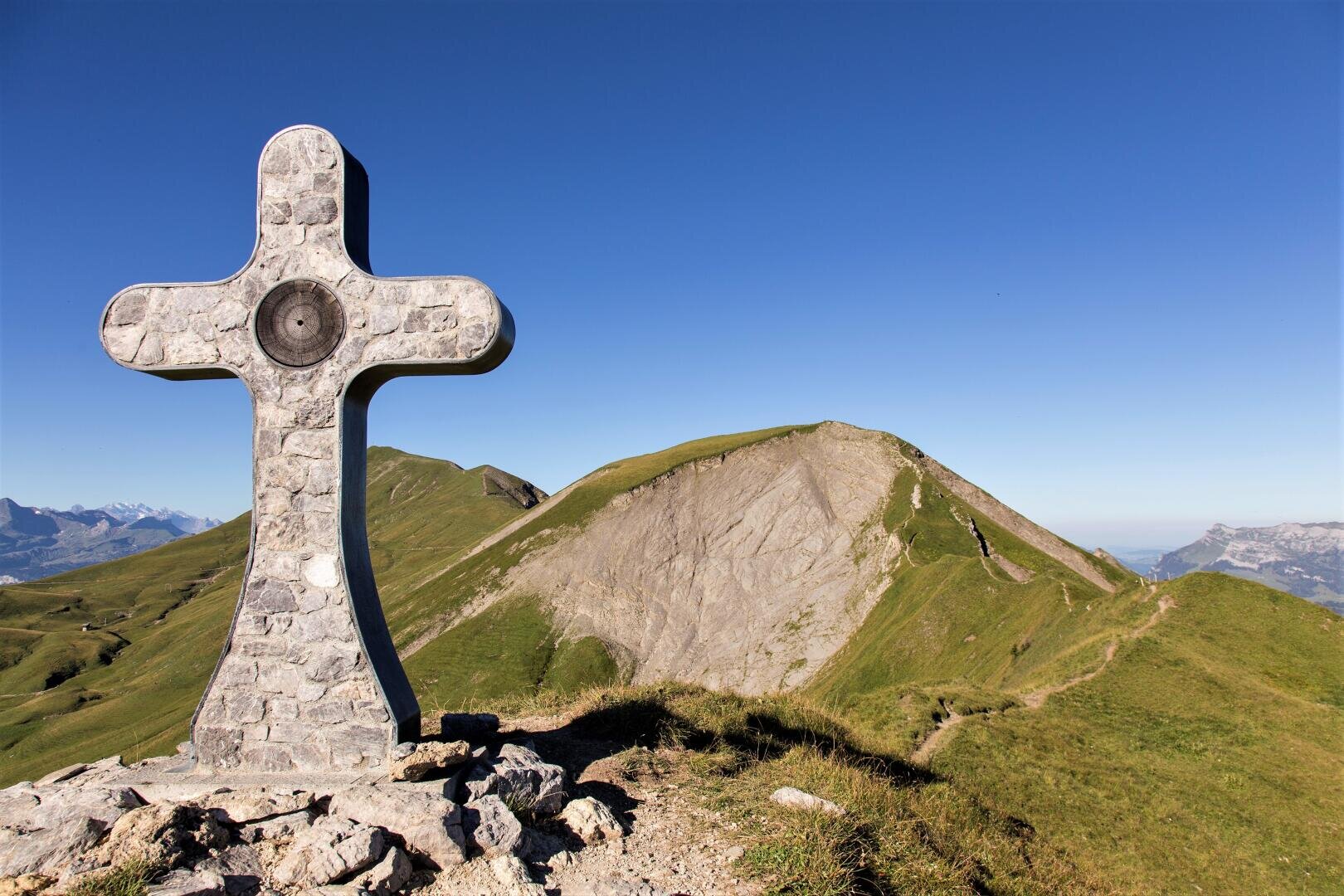 Ein steinernes Kreuzdenkmal steht auf einem grasbewachsenen Bergrücken unter strahlend blauem Himmel, und verschlungene Pfade führen hinauf zu den nahe gelegenen Gipfeln.