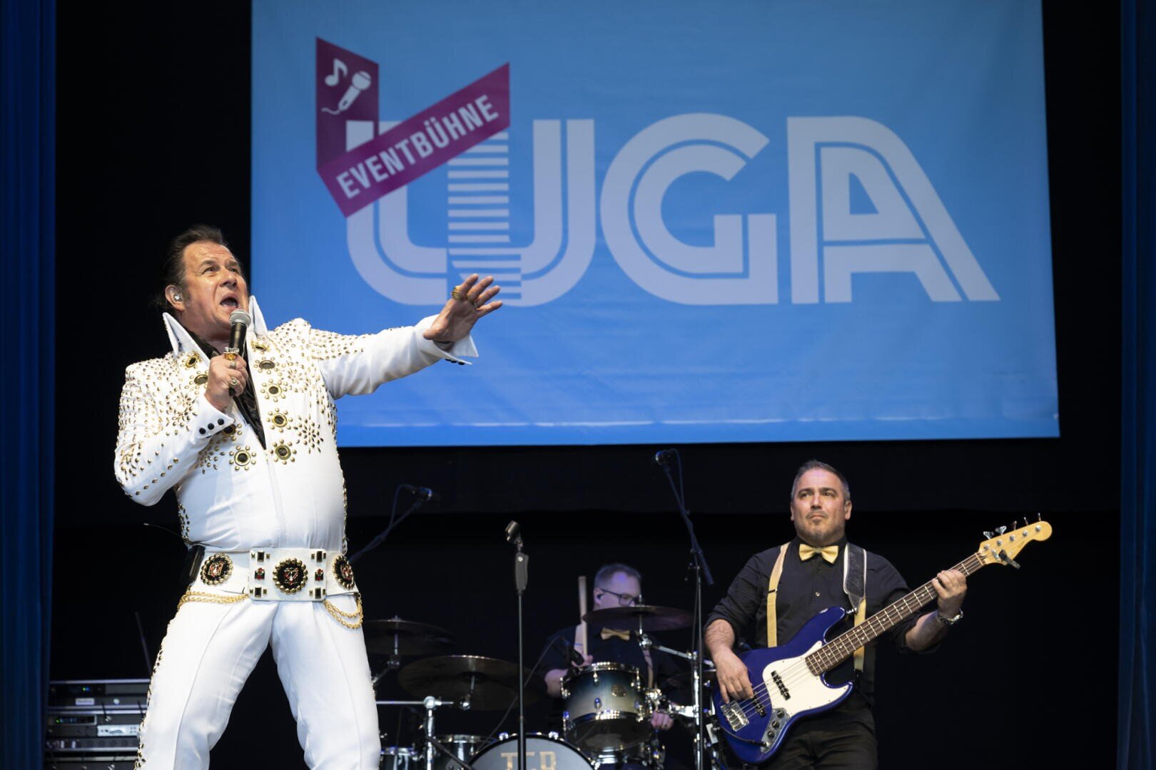 Ein als Elvis Presley verkleideter Darsteller singt leidenschaftlich auf der Bühne mit einer Live-Band. Hinter ihnen steht ein großes blaues Schild mit der Aufschrift UGA EVENTBÜHNE. Ein Bassist und ein Schlagzeuger sind ebenfalls zu sehen.