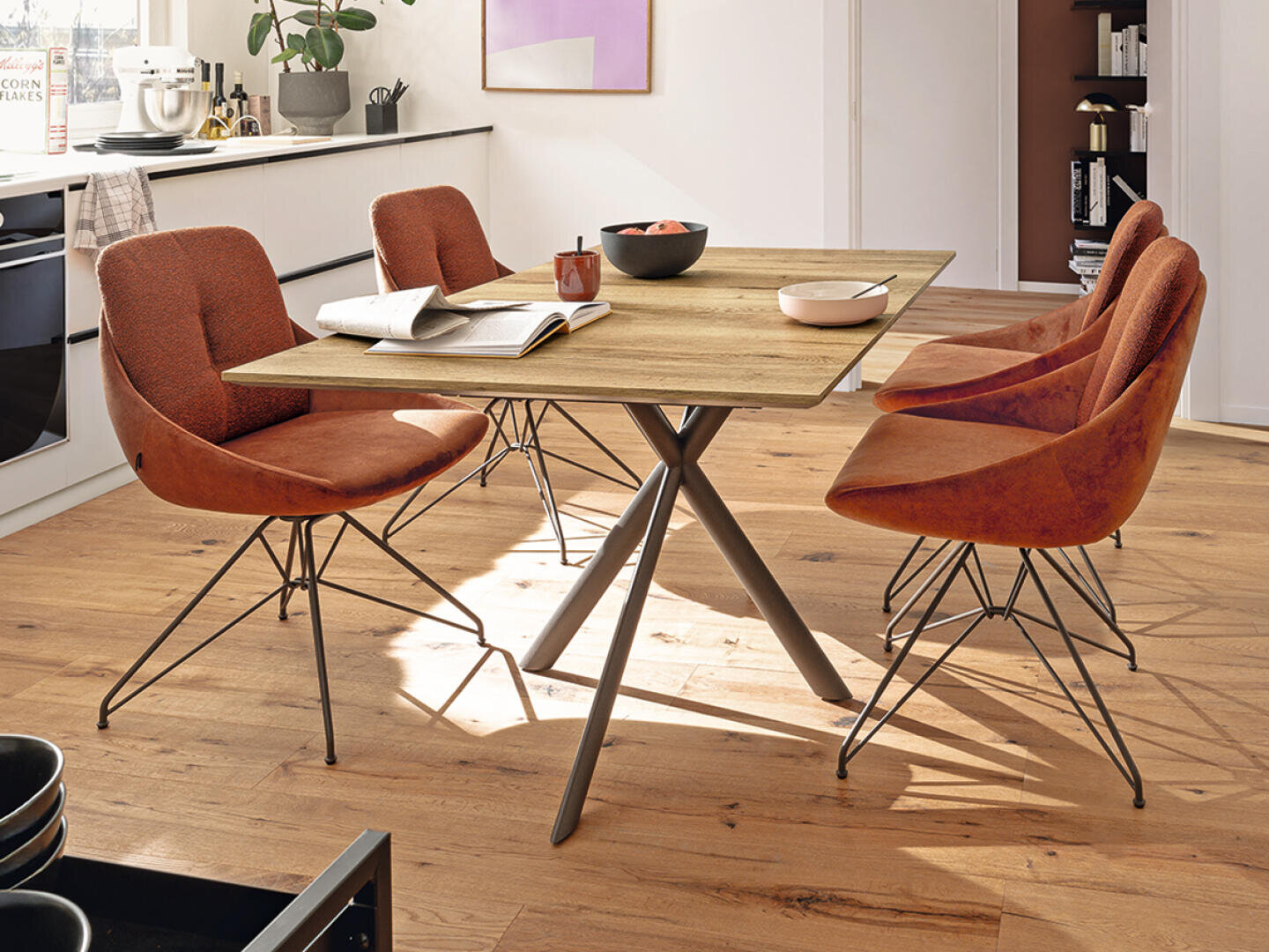 Ein moderner Essbereich mit einem Holztisch, vier orangefarbenen gepolsterten Stühlen, einem aufgeschlagenen Buch und Geschirr auf dem Tisch. Der Raum ist mit Holzfußboden und einer hellen Küche im Hintergrund ausgestattet.