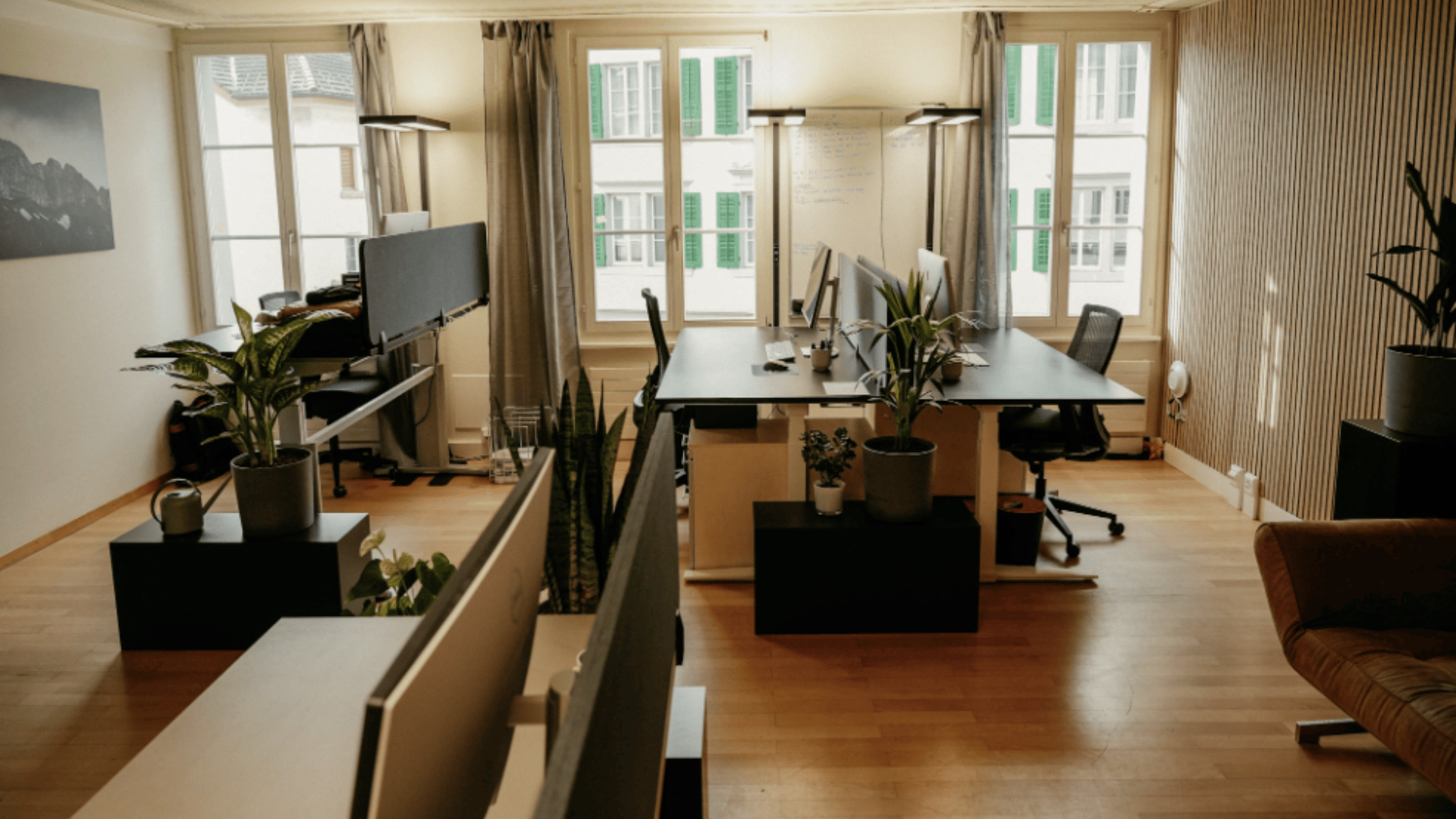 Ein helles, modernes Büro mit Holzböden, Schreibtischen mit Computern, Topfpflanzen und großen Fenstern, durch die natürliches Licht einfällt. Um die Schreibtische herum stehen Stühle, und draußen sind die Gebäude der Stadt zu sehen.