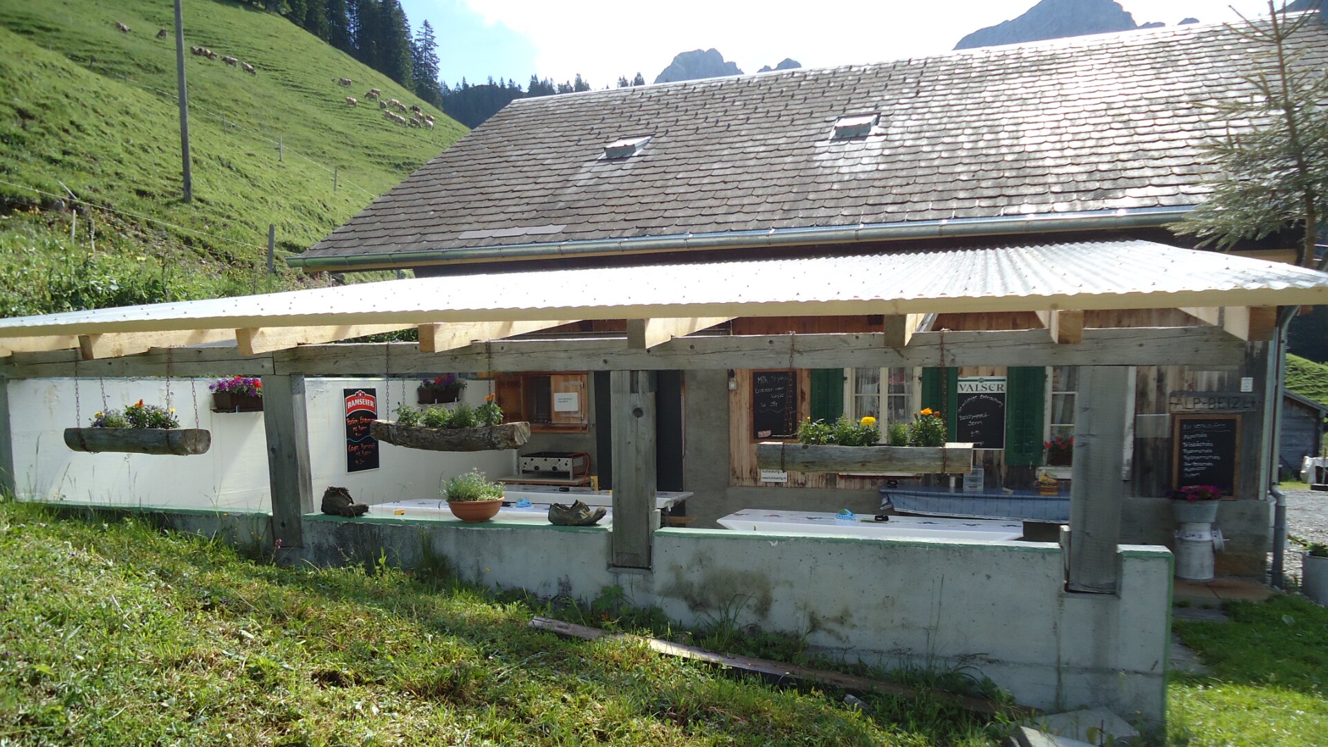 Ein rustikales Gebäude mit schrägem Dach und einem überdachten Außenbereich mit Topfpflanzen, Blumen und Speisekarten auf Kreidetafeln an der Wand; im Hintergrund sind grüne Hügel und Berge zu sehen.