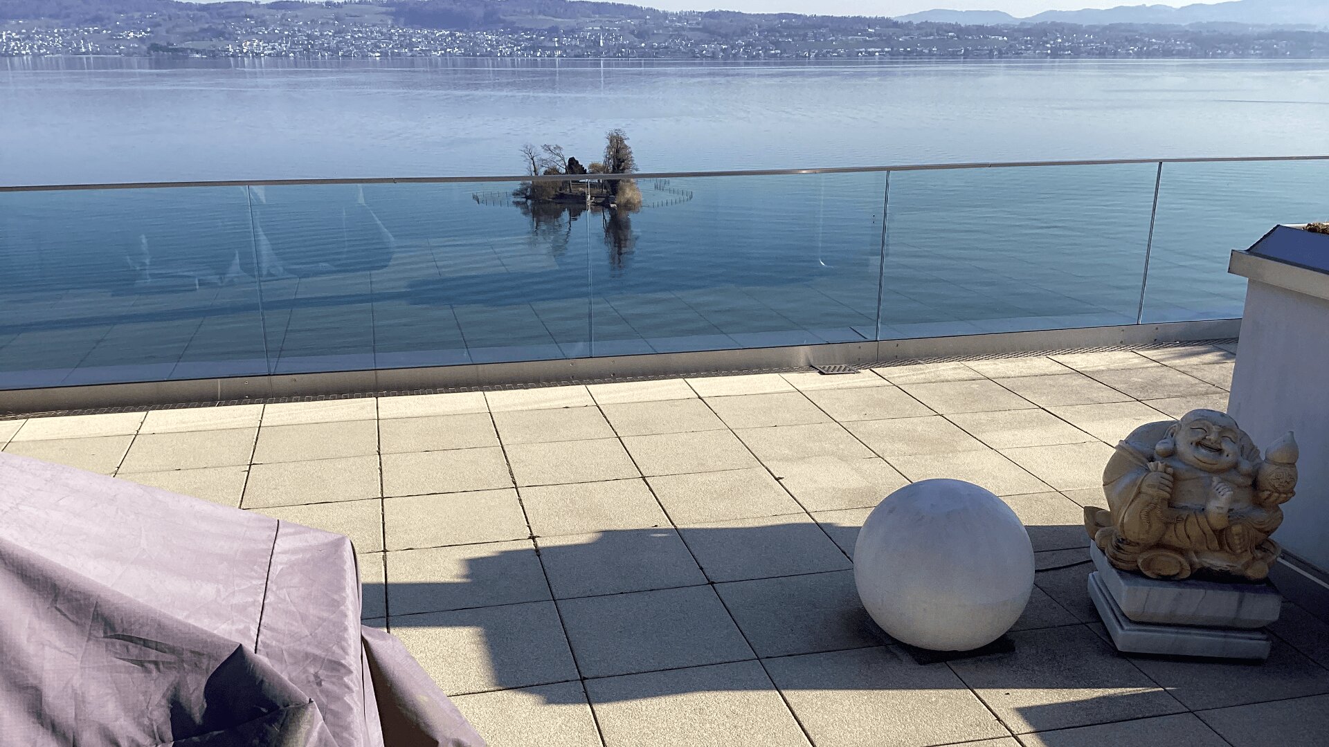 Eine Seeterrasse mit hellgrauen Fliesen, einer kugelförmigen Steinskulptur, einer lachenden Buddha-Statue und einem Glasgeländer mit Blick auf ruhiges blaues Wasser und eine kleine Insel, mit Hügeln und einer Stadt im Hintergrund.