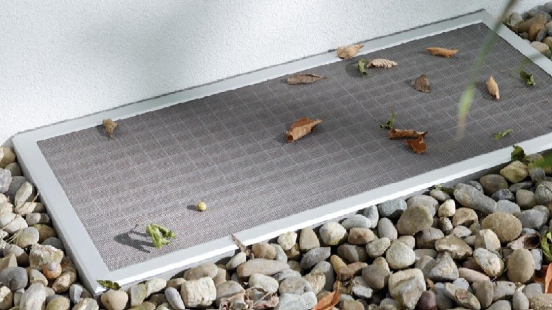 Ein mit einem Gitter abgedeckter Kellerfensterschacht, umgeben von kleinen Steinen und einigen getrockneten Blättern, die auf dem Gitter verstreut sind.