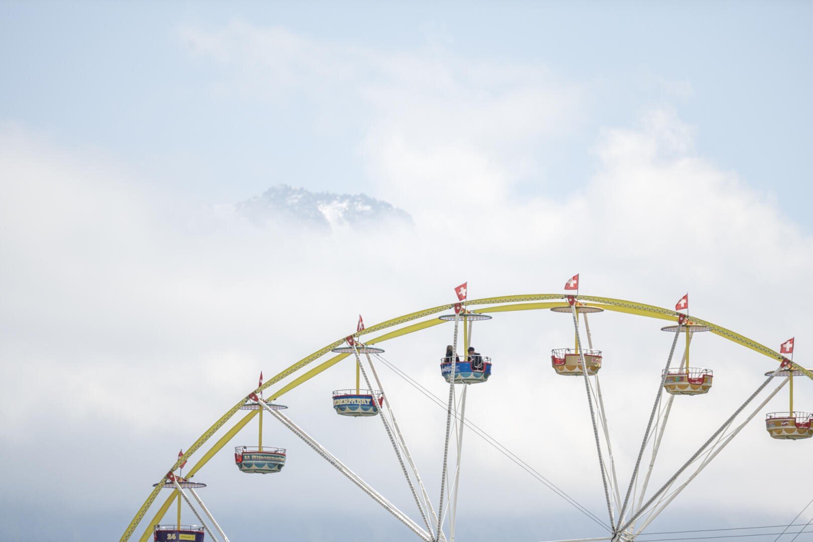 Ein teilweise sichtbares Riesenrad mit bunten Gondeln und kleinen roten Fahnen steht vor einem wolkenverhangenen Himmel, und im Hintergrund ist schwach ein schneebedeckter Berggipfel zu erkennen.