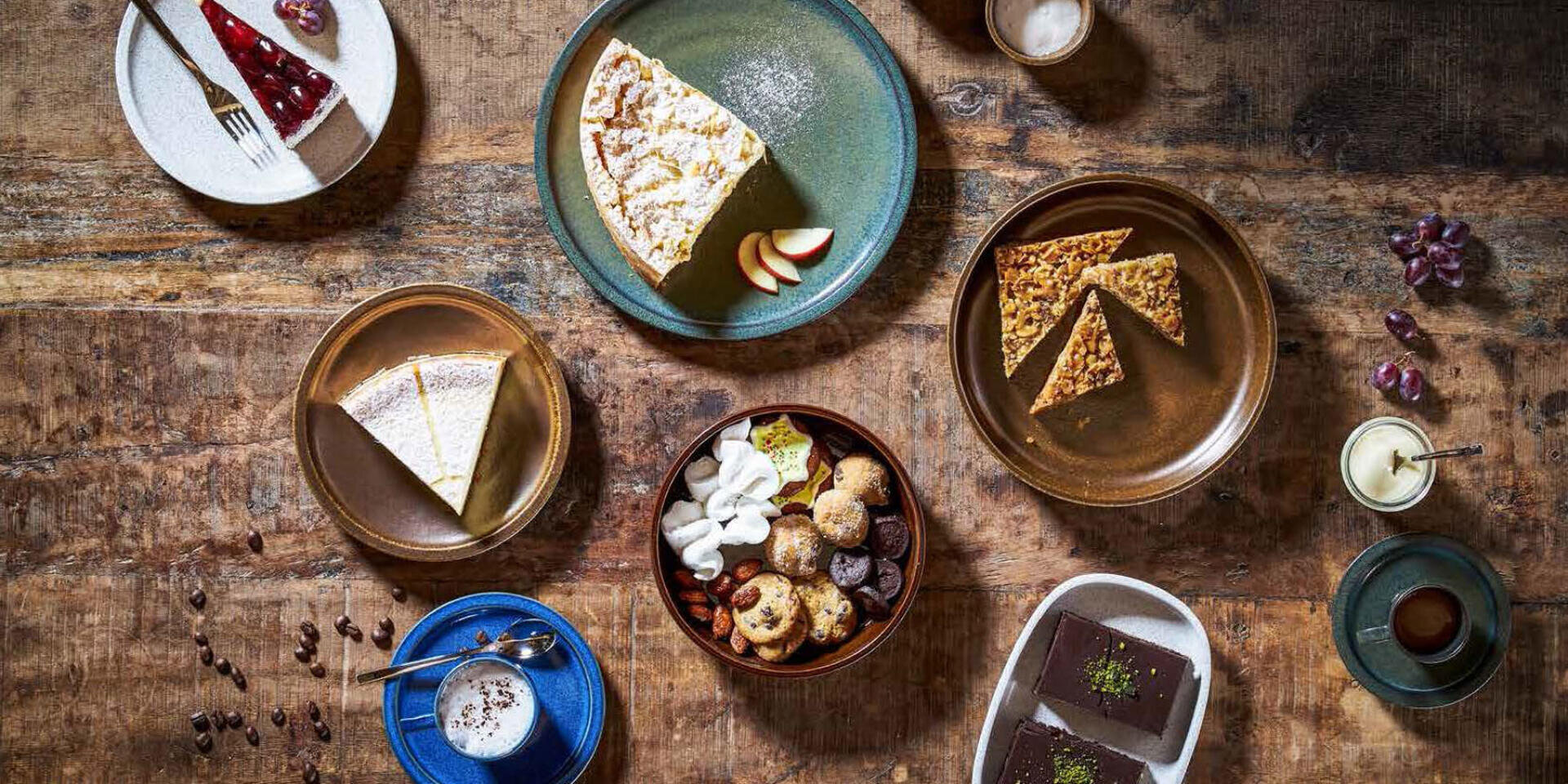 Ein rustikaler Holztisch mit verschiedenen Desserts, darunter Kuchenstücke, Kekse, Brownies und Getränke, die kunstvoll auf Keramiktellern und -schalen angerichtet sind.