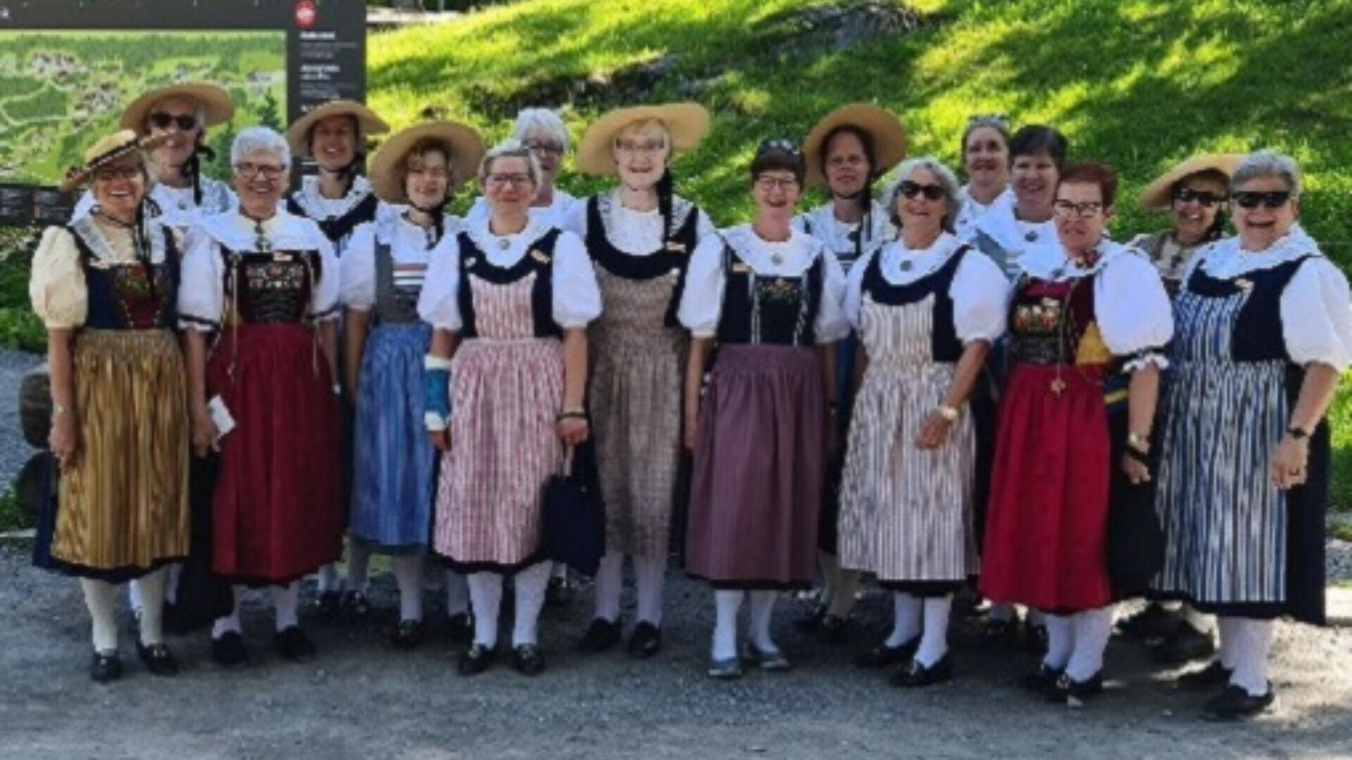 Eine Gruppe von Frauen steht im Freien, lächelnd und in traditioneller europäischer Volkstracht mit Schürzen, Blusen und Hauben. Im Hintergrund sind grünes Gras und ein Landkartenschild zu sehen.