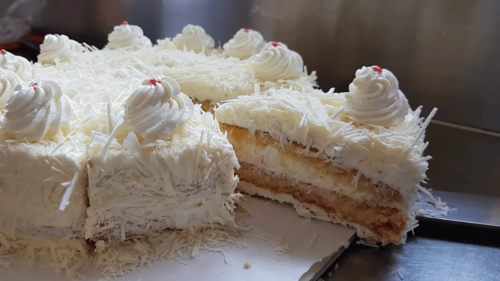 Ein in Scheiben geschnittener weißer Waldkuchen mit Schlagsahne und roten Kirschen, bedeckt mit weißen Schokoladensplittern, liegt auf einer flachen Oberfläche, von der ein Stück entfernt wurde.