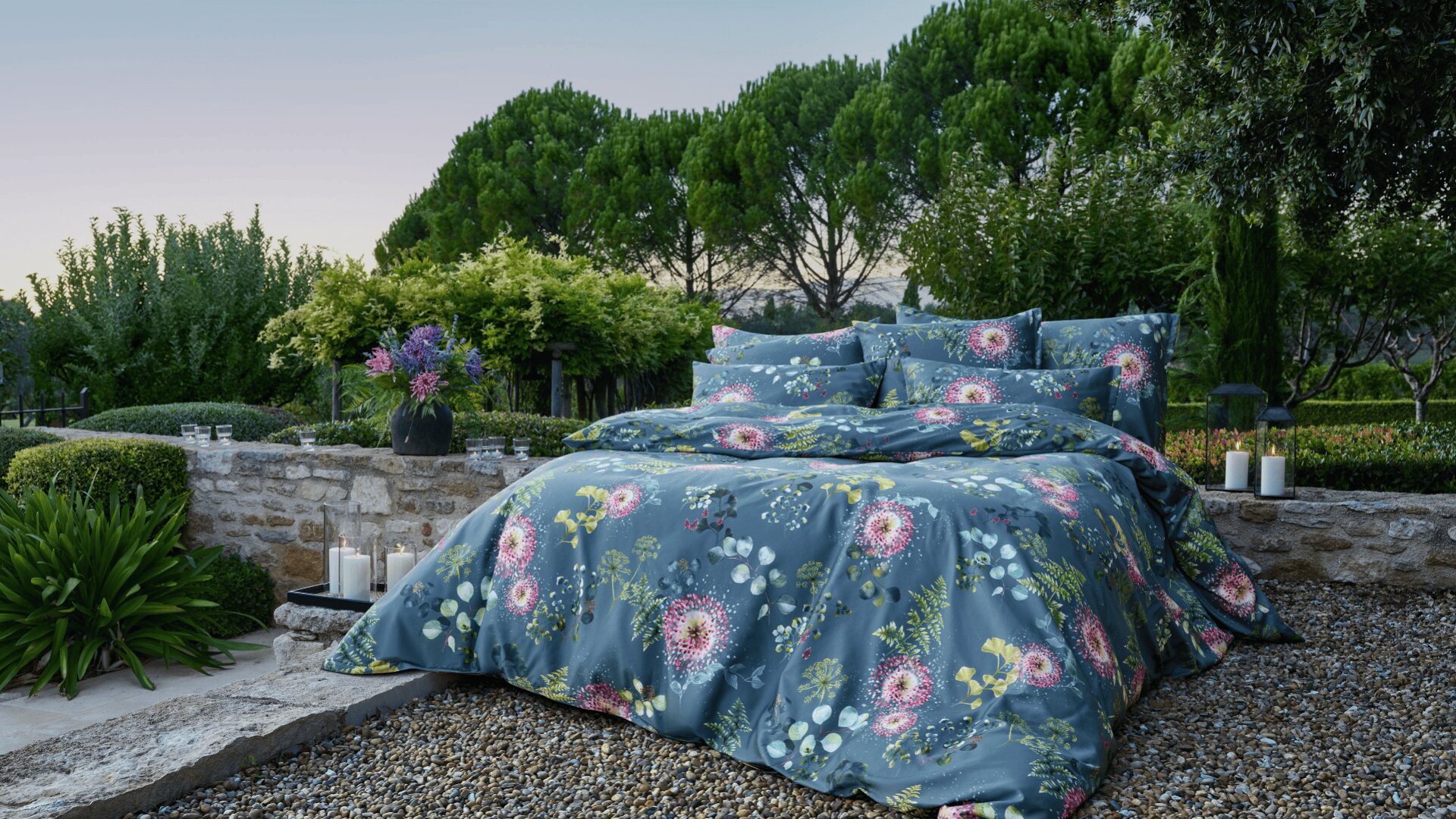Ein ordentlich gemachtes Bett mit blauer Blumenbettwäsche steht im Freien auf Kies, umgeben von üppigem Grün, Steinmauern, Kerzen und einer Blumenvase, mit hohen Bäumen im Hintergrund in der Abenddämmerung.
