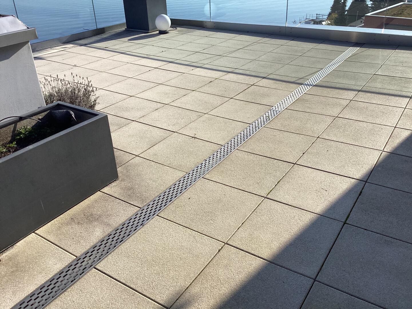 Eine Dachterrasse mit großen hellen Fliesen, einem diagonal verlaufenden Entwässerungsgitter, einem rechteckigen Pflanzgefäß mit spärlicher Bepflanzung, einem Glasgeländer und einem Blick auf das Wasser im Hintergrund.
