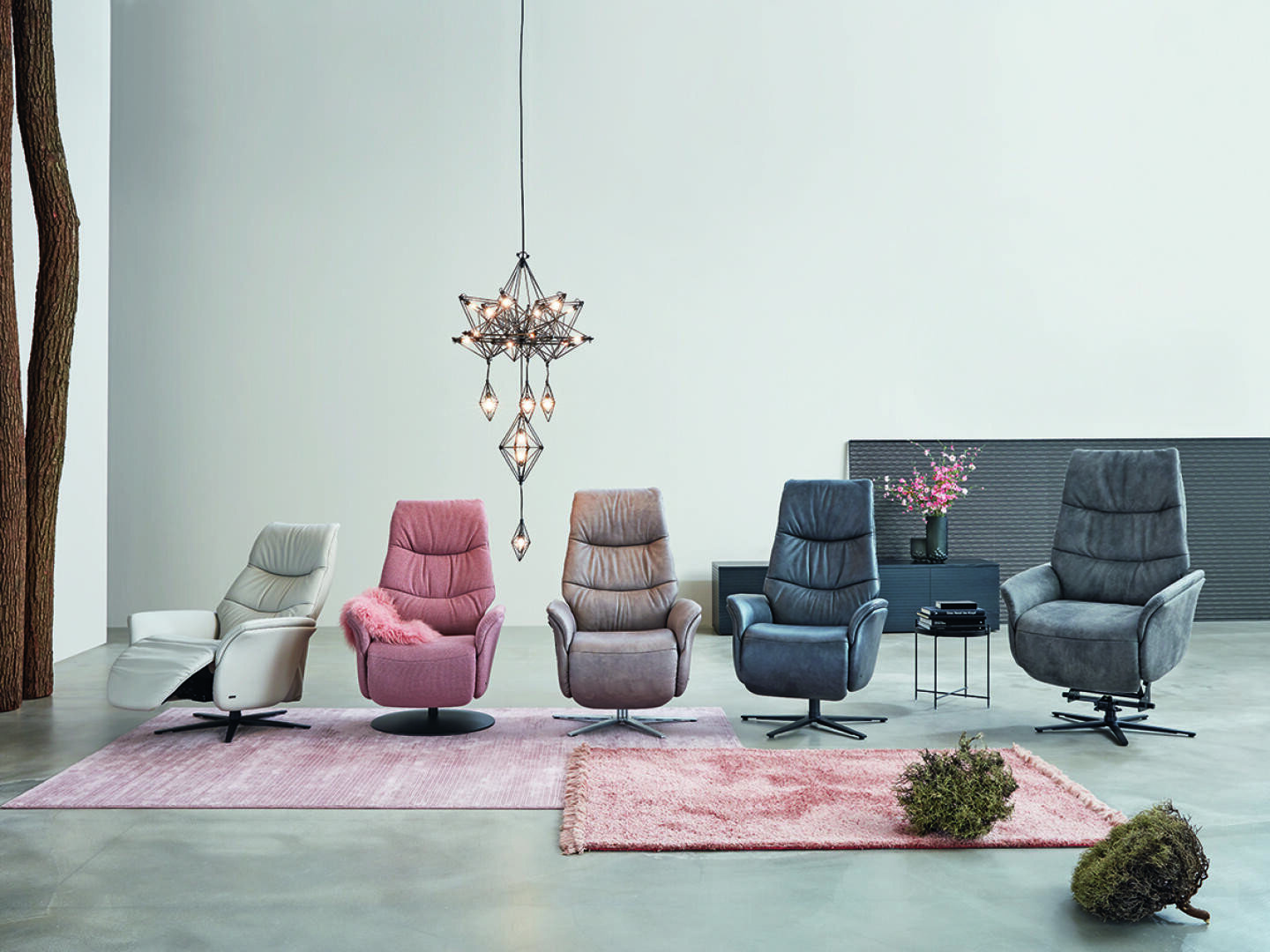 Fünf moderne Sessel in verschiedenen Farben (weiß, rosa, taupe, dunkelblau und grau) stehen auf rosa Teppichen unter einem geometrischen Kronleuchter in einem minimalistischen Raum mit einem Beistelltisch, Blumen und einem Baumstamm.