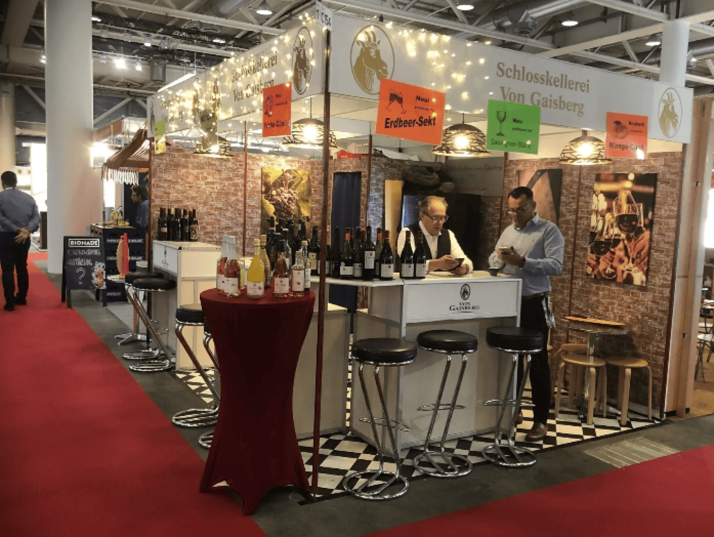 Ein Weinstand bei einer Indoor-Veranstaltung mit Flaschen auf einer Theke, zwei Männern, die dahinter stehen, und bunten Schildern, auf denen verschiedene Getränke angeboten werden. Der Stand ist mit Lichtern und einer Backsteinmauer als Hintergrund dekoriert.