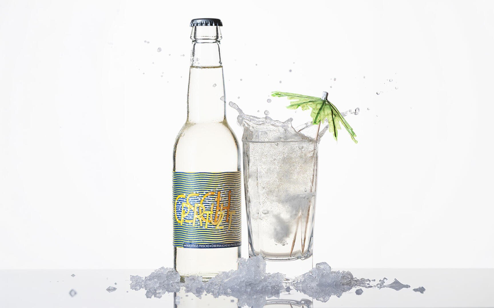 Eine klare Getränkeflasche mit der Aufschrift OSHU steht neben einem mit Eis gefüllten Glas, einer klaren Flüssigkeit und einem grünen Cocktailschirmchen. Eiswürfel und Wasserspritzer umgeben beide auf einer spiegelnden Oberfläche.