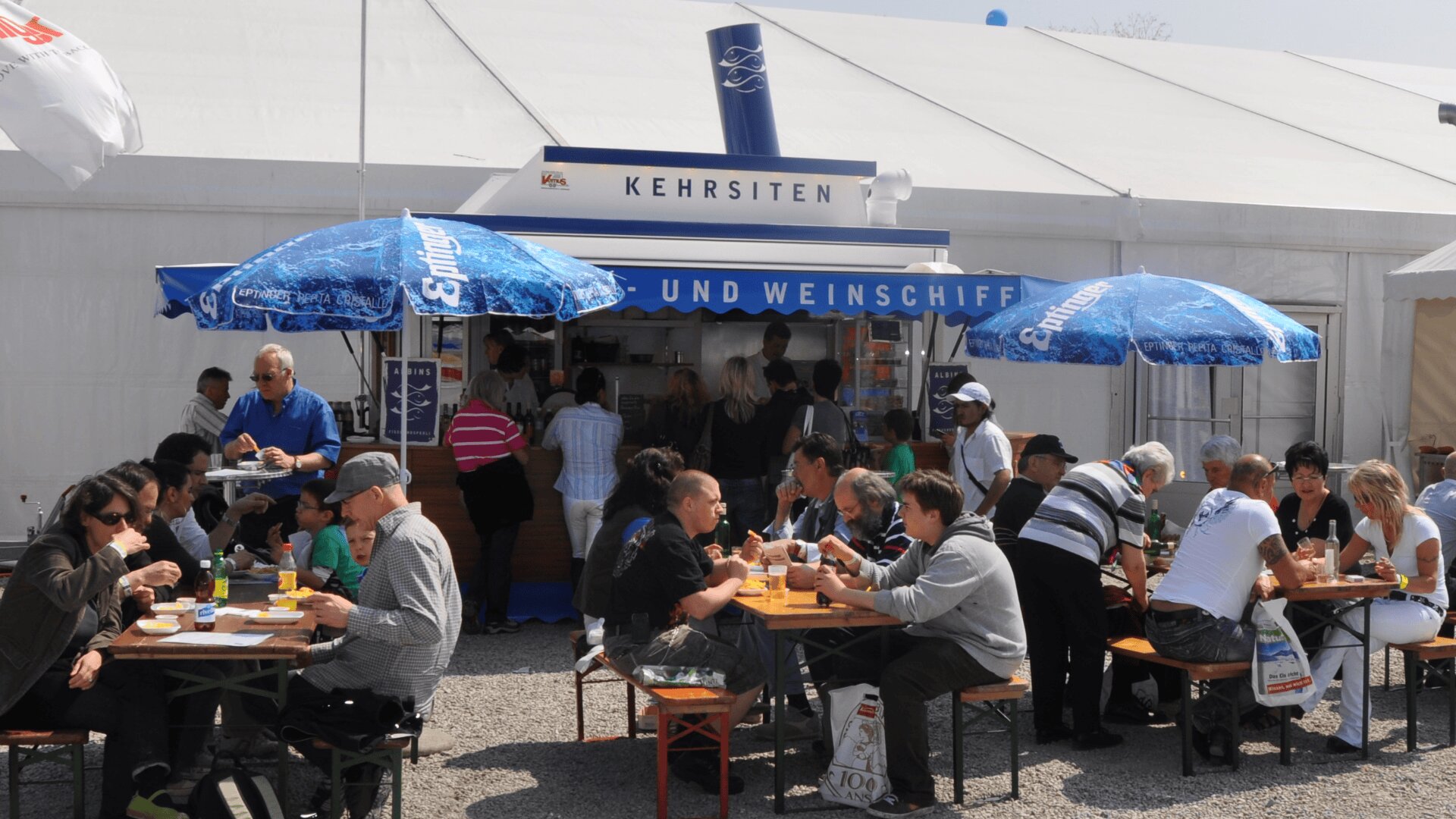 Menschen sitzen an Tischen im Freien unter blauen Sonnenschirmen, essen und trinken vor einem Essens- und Weinstand mit der Aufschrift Kehrsiten und einem weißen Zelt im Hintergrund. Die Szene ist lebhaft und erinnert an ein Fest oder einen Jahrmarkt.