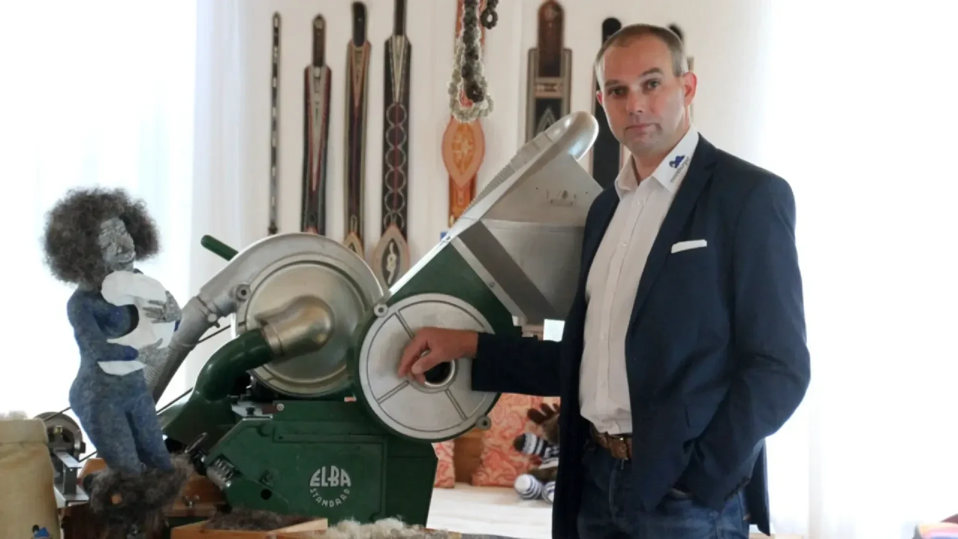 Ein Mann im Anzug steht neben einer alten grünen Elba-Maschine in einem Raum, der mit hängenden Gürteln und Kunsthandwerk dekoriert ist. Er hat eine Hand auf der Maschine und blickt in die Kamera.