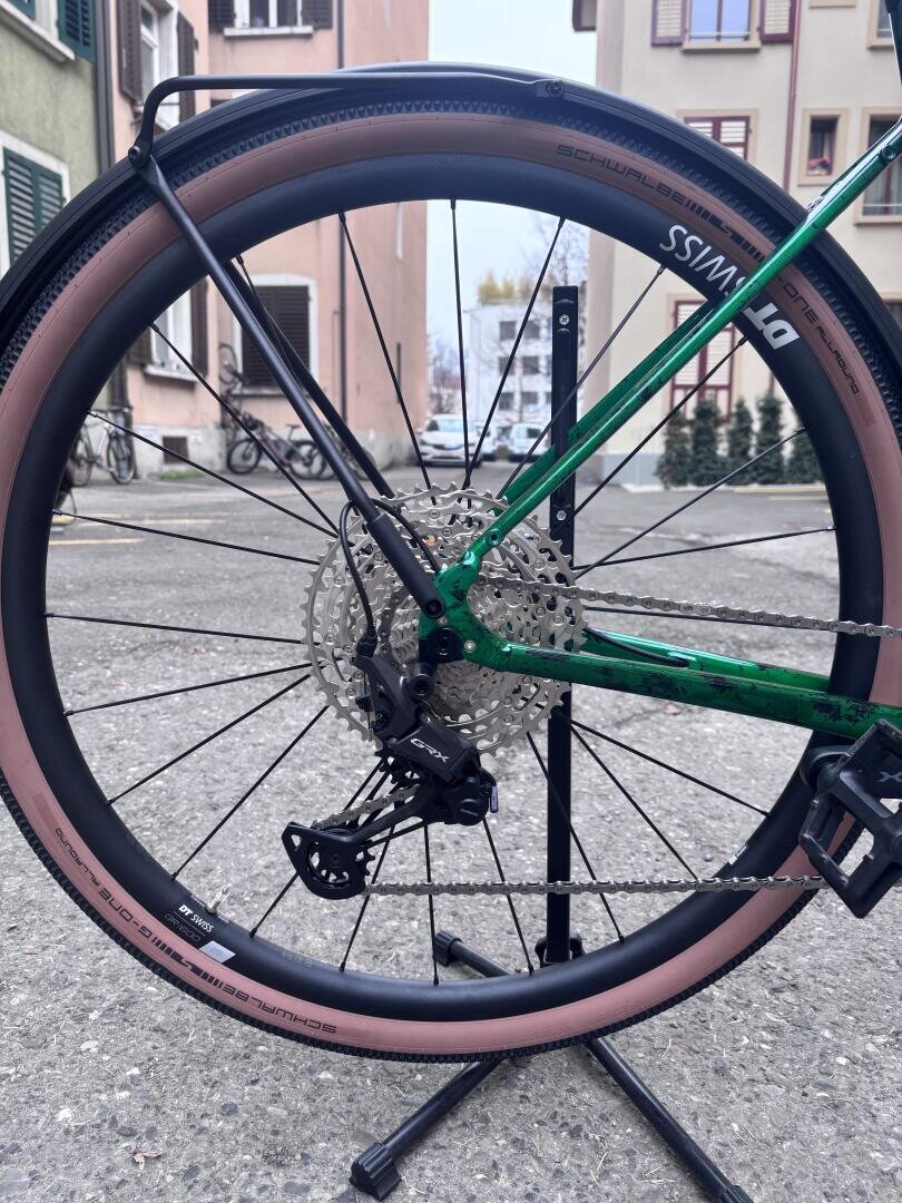 Nahaufnahme des Hinterrads und der Gangschaltung eines grünen Fahrrads mit rosa Reifen, das auf einem Ständer in einer städtischen Umgebung mit Wohnhäusern und anderen Fahrrädern im Hintergrund abgestellt ist.