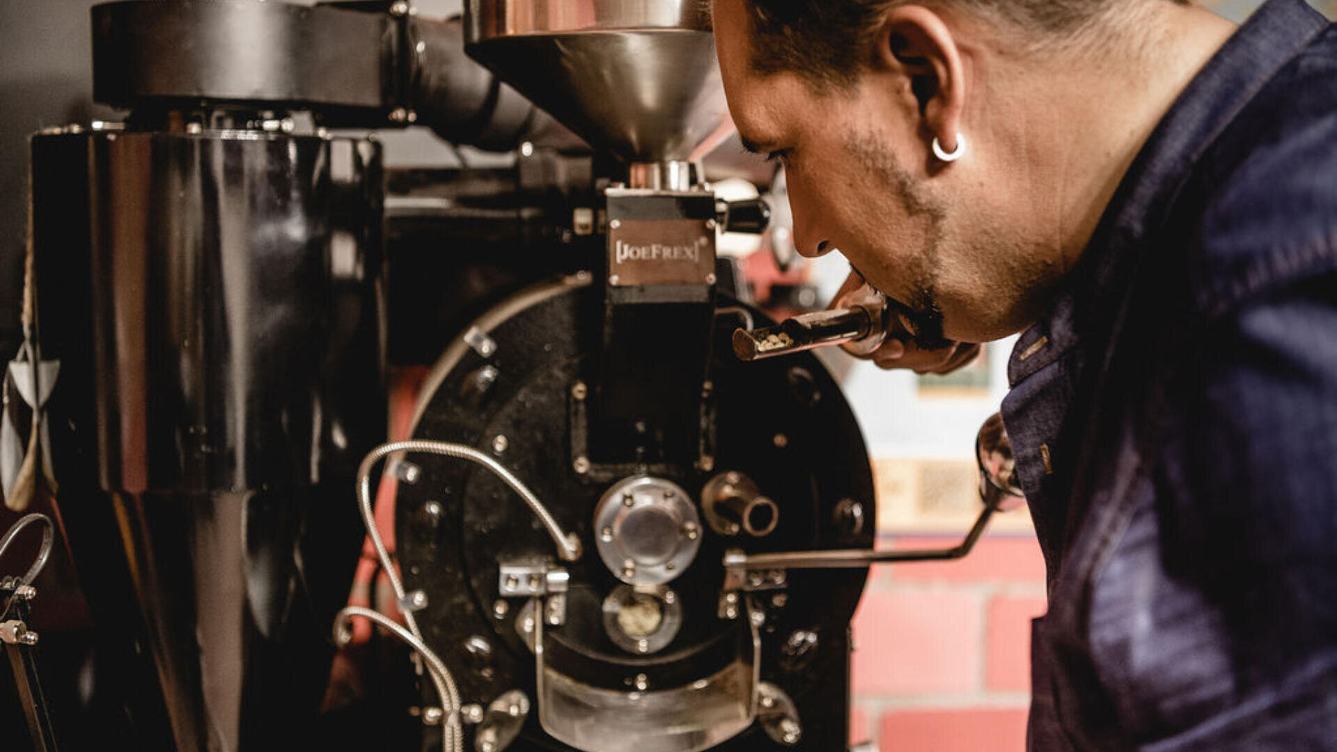 Ein Mann in Jeansjacke prüft eine Probe aus einer Kaffeeröstmaschine in einer Industrieumgebung mit freiliegenden Ziegelwänden genau.