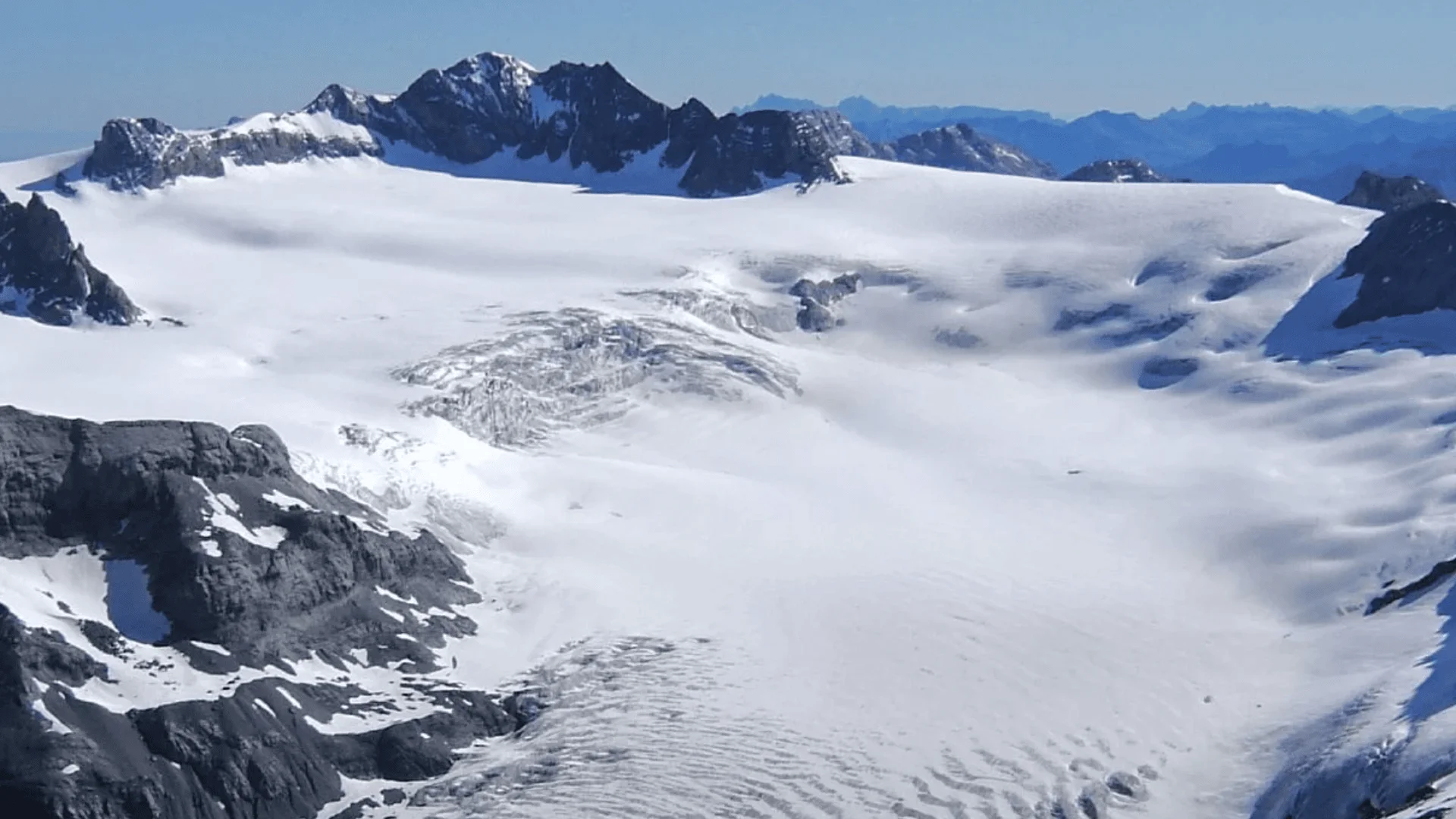 Schneebedeckte Berge und ein Gletscher unter strahlend blauem Himmel, mit felsigen Gipfeln und Graten, die sich über die weite Eislandschaft erheben.