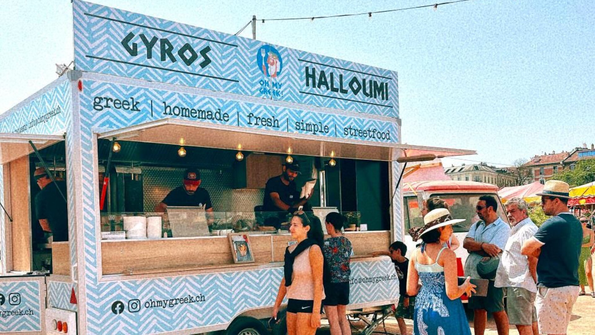 An einem sonnigen Tag stehen die Leute vor einem blau gemusterten griechischen Imbisswagen an, der Gyros und Halloumi verkauft. Das Personal bereitet das Essen drinnen zu, und die Szene hat eine entspannte, sommerliche Atmosphäre im Freien.