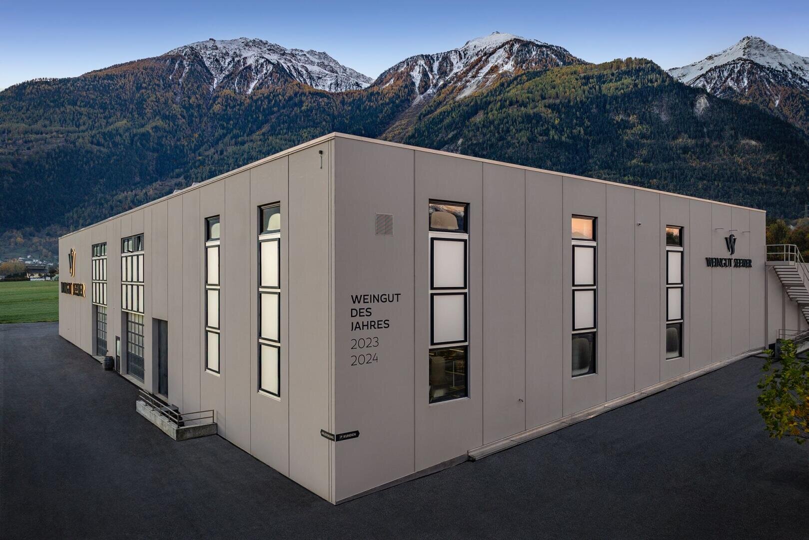 Ein modernes, helles Gebäude mit großen Fenstern steht vor einem baumbewachsenen Gebirge mit schneebedeckten Gipfeln. An der Wand des Gebäudes ist die Aufschrift "Weingut des Jahres 2023 2024" zu lesen.