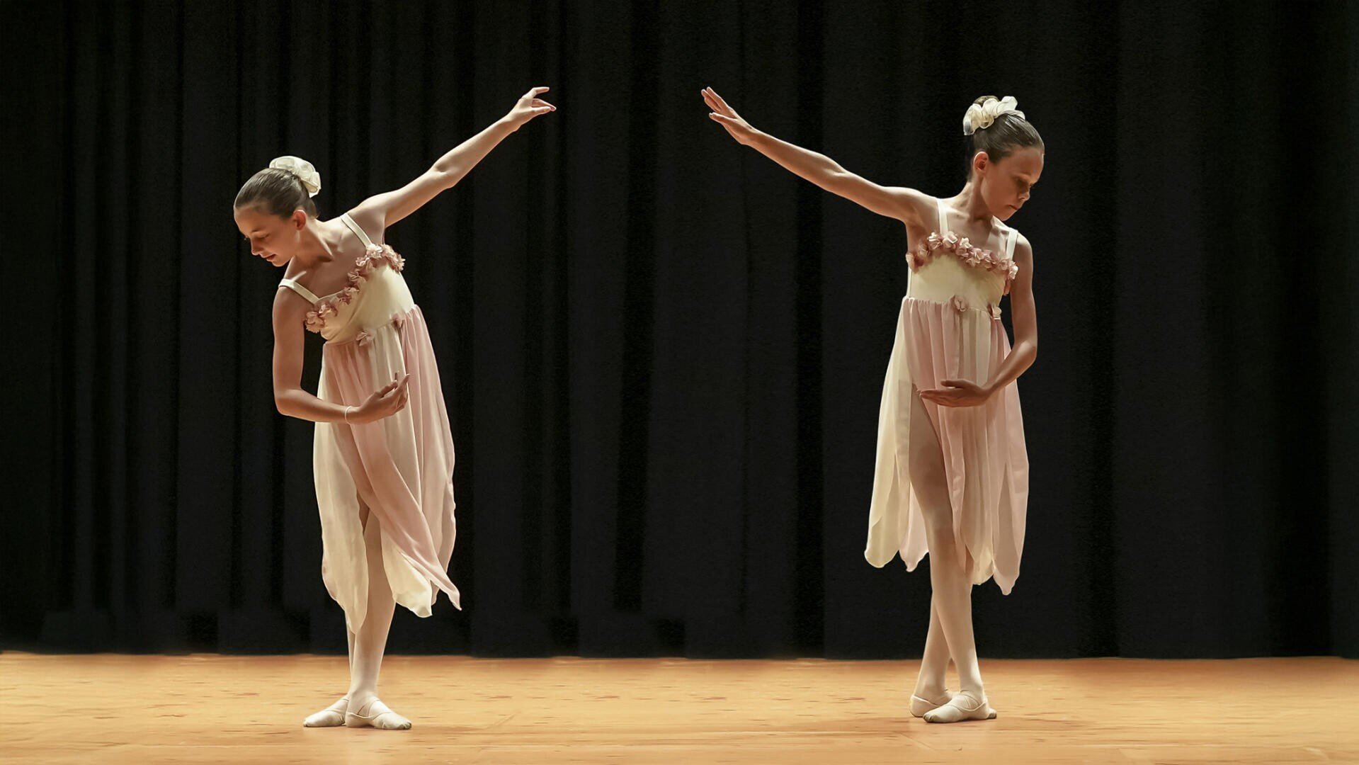Zwei junge Balletttänzerinnen in passenden rosafarbenen Kleidern und weißen Ballettschuhen posieren anmutig auf der Bühne, jede mit einem ausgestreckten Arm und gesenktem Kopf, vor einem schwarzen Vorhang.