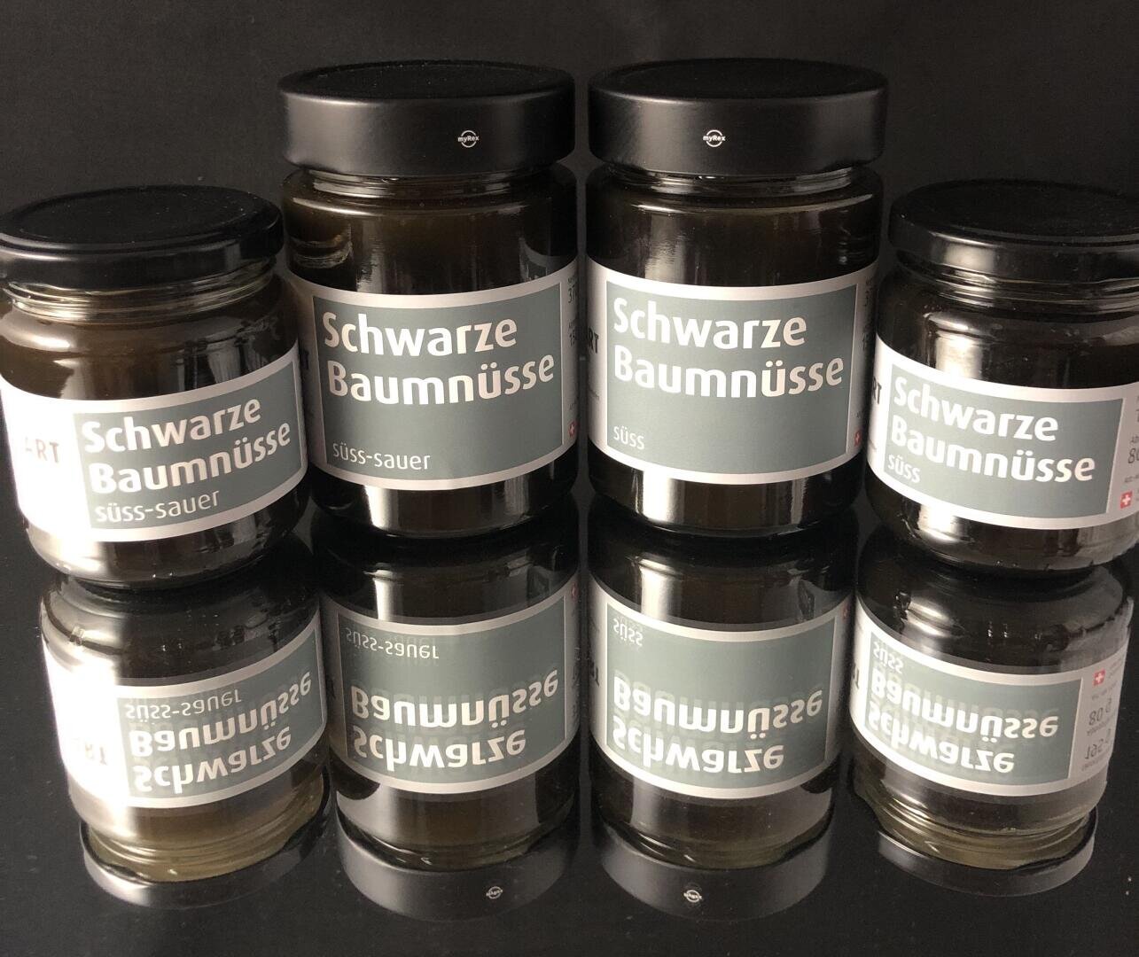 Sechs Gläser mit der Aufschrift Schwarze Baumnüsse sind in zwei Reihen angeordnet und spiegeln sich auf einer glänzenden schwarzen Oberfläche. Auf den Etiketten steht außerdem "süss-sauer".