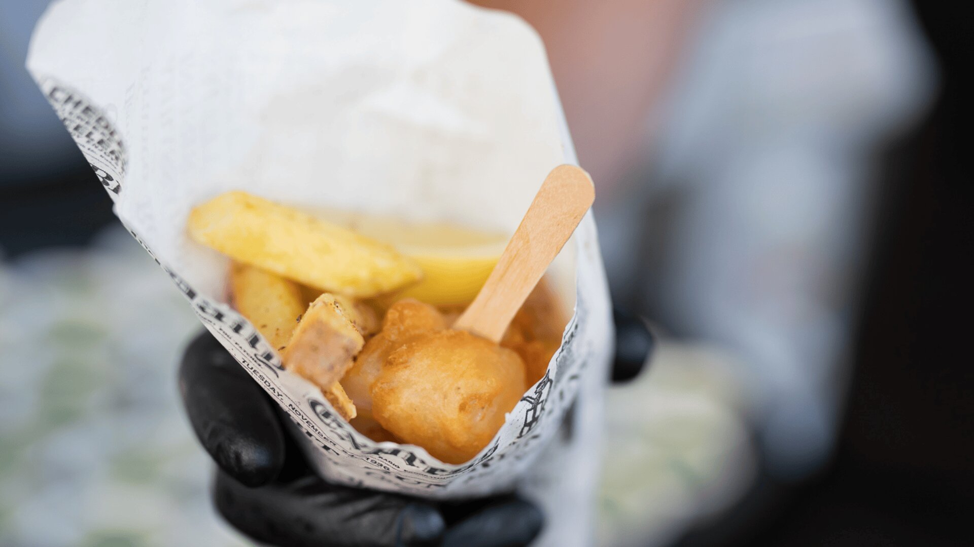 Eine behandschuhte Hand hält eine Papiertüte, die mit frittiertem Essen gefüllt ist, unter anderem mit panierten Gegenständen und Kartoffelecken, und einen Holzstab zum Essen.