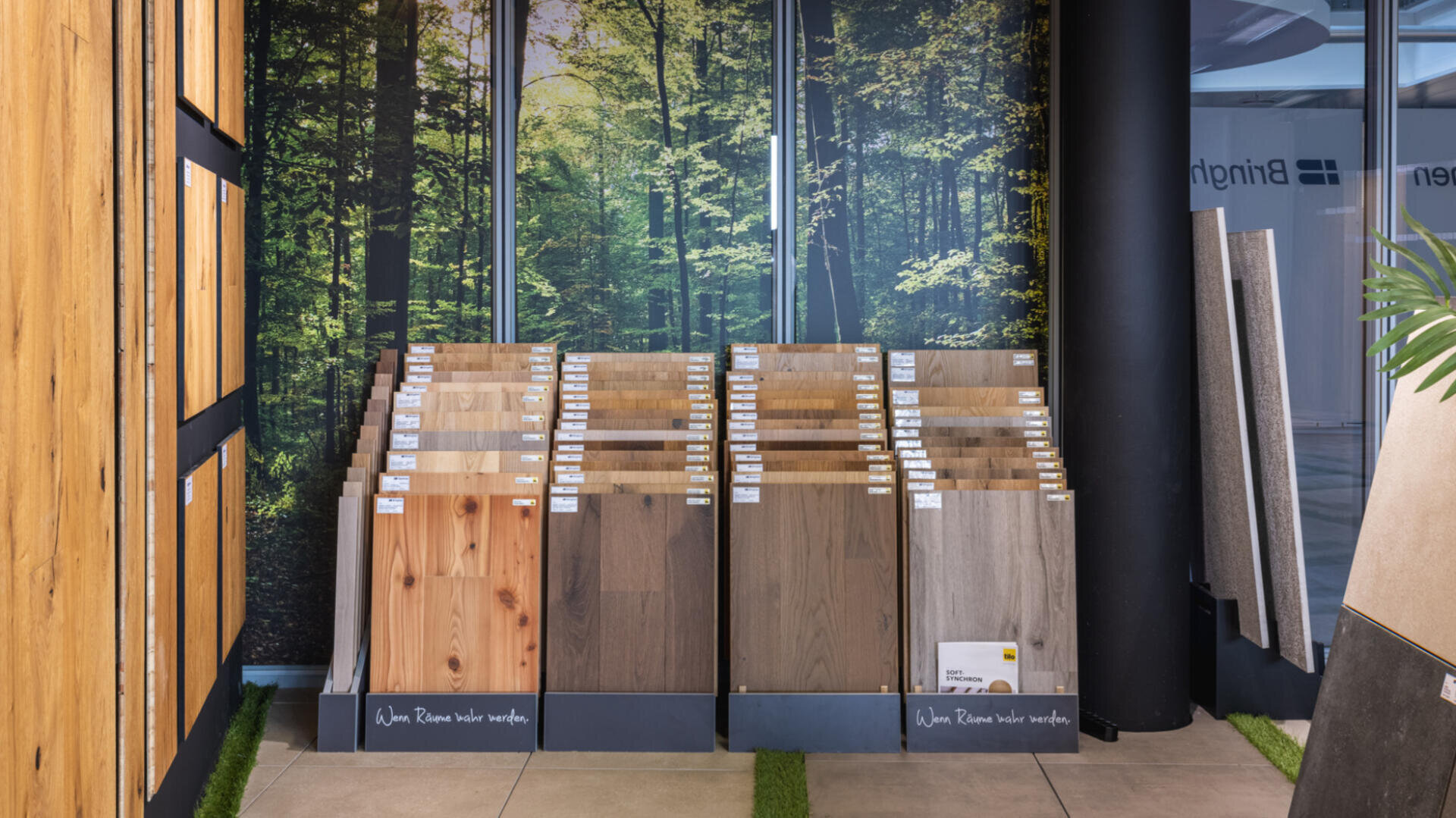 Ausstellung verschiedener Holz- und Laminatfußbodenmuster in Reihen in einem Ausstellungsraum, mit einem großen Wandbild eines sonnenbeschienenen Waldes im Hintergrund.