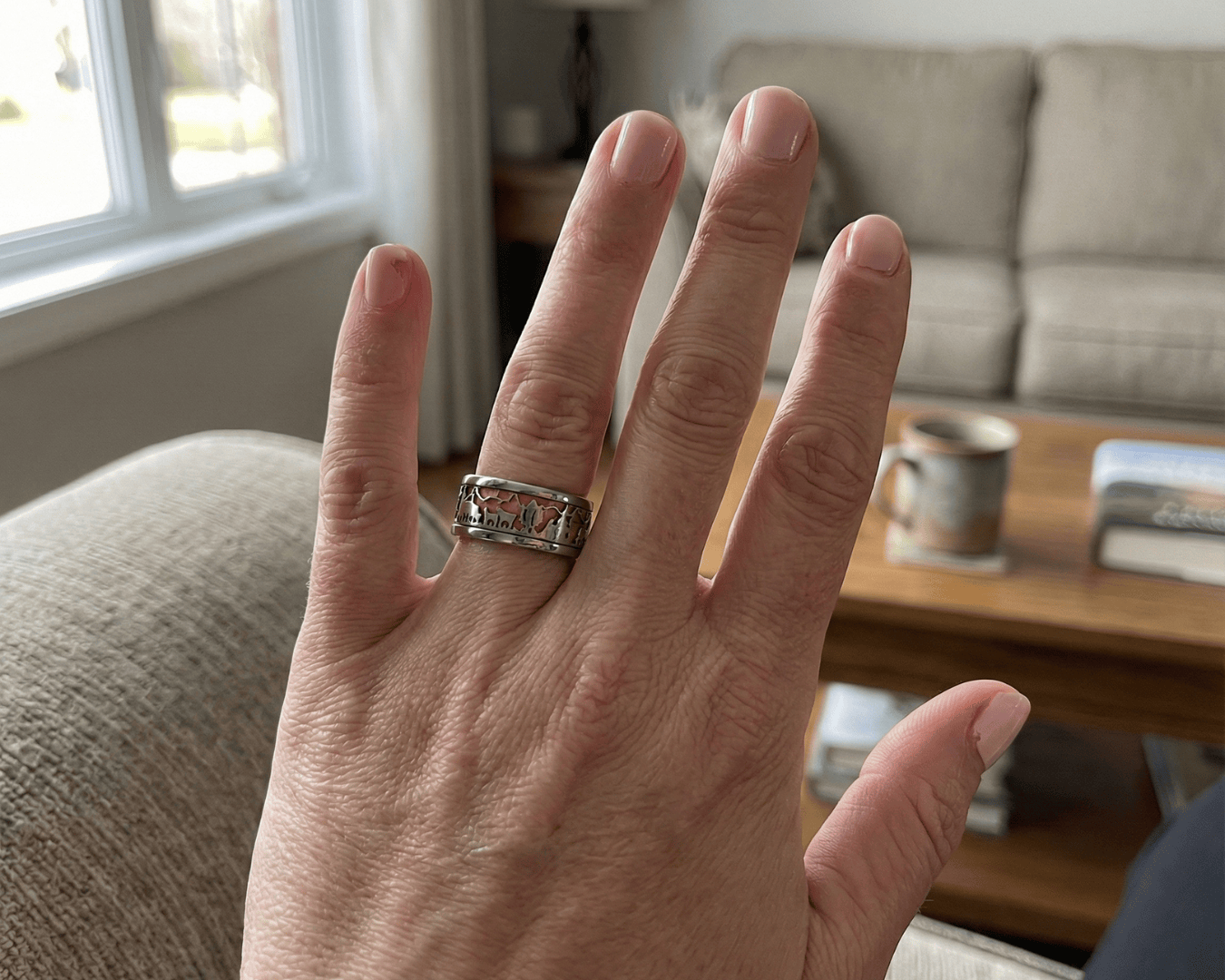 Eine Hand mit sauber geschnittenen Fingernägeln und einem silbernen Ring wird in einem Haus hochgehalten. Im Hintergrund stehen eine beigefarbene Couch, ein Couchtisch mit einem Becher und einige Bücher. Das Tageslicht fällt durch ein Fenster.