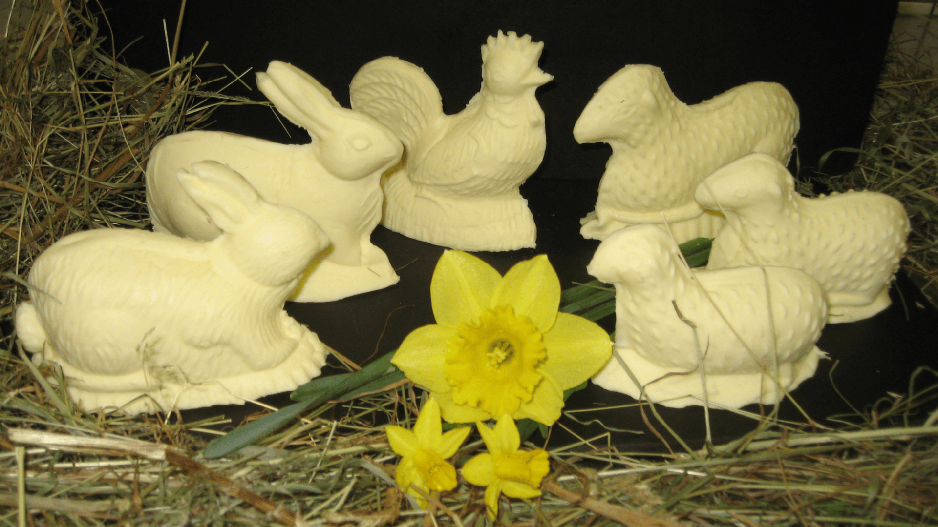 Osterskulpturen aus Butter in Form von Hasen, Schafen und einem Hahn, umgeben von Heu und gelben Osterglocken.