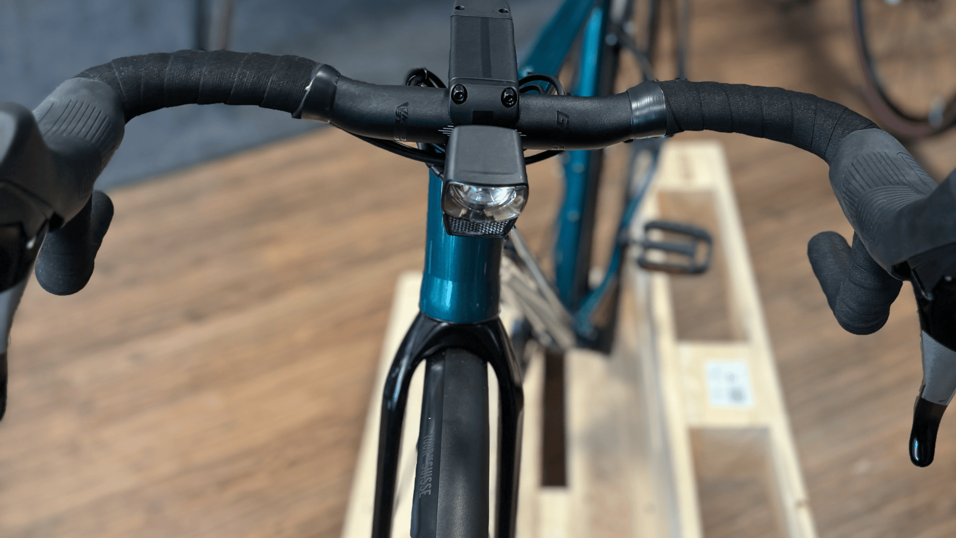 Blick von oben auf einen Fahrradlenker, einen Frontscheinwerfer und das Vorderrad, wobei das Fahrrad auf einem hölzernen Ausstellungsständer in einem Innenraum steht.