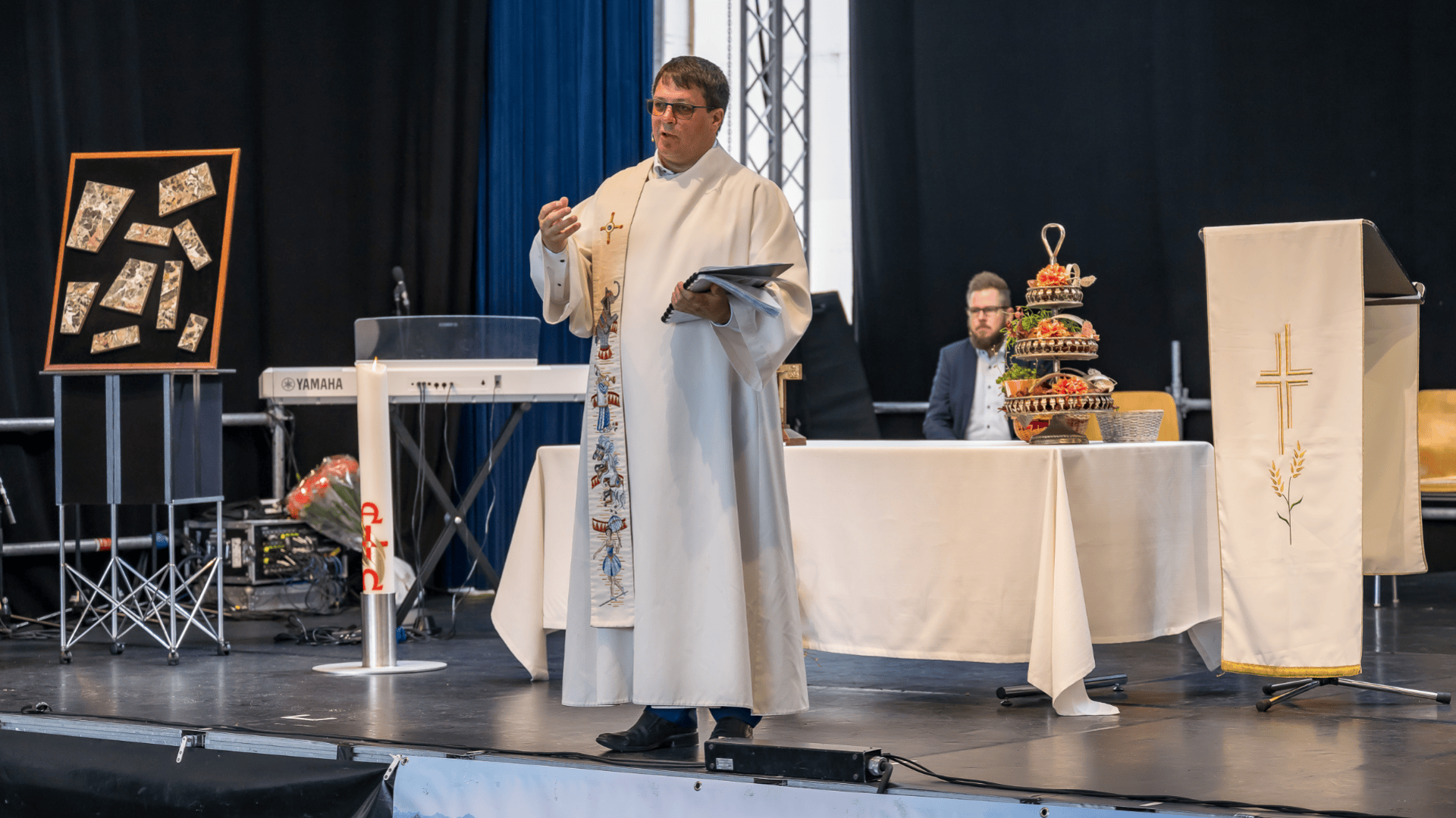 Ein Priester in weißem Gewand spricht auf der Bühne und hält ein Buch in der Hand. Hinter ihm sitzt ein Mann an einem Tisch mit einem Blumengesteck und einem großen verzierten Brot. Im Hintergrund sind eine Tastatur, gerahmte Bilder und religiöse Symbole zu sehen.
